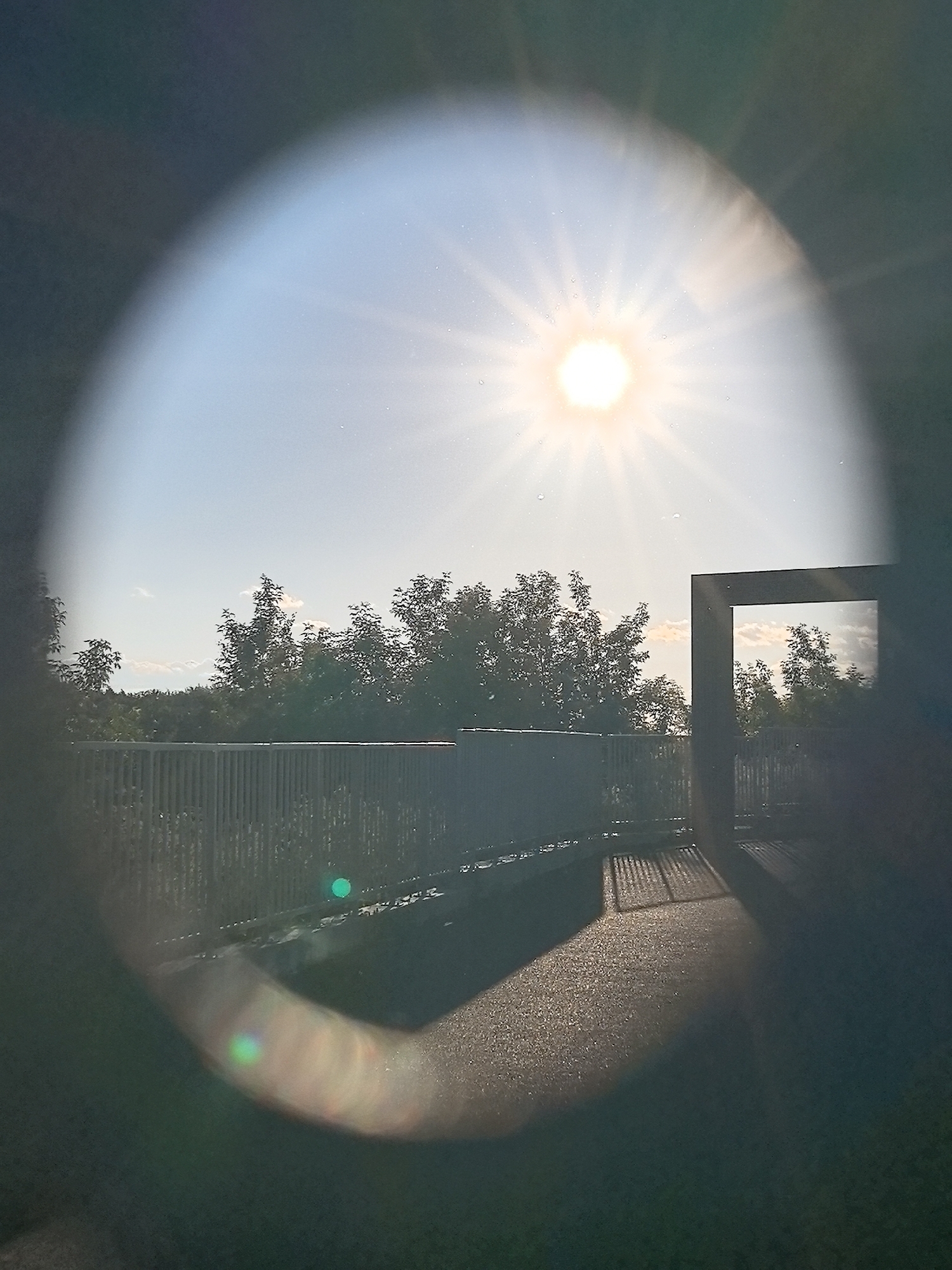 The image shows a view through an oval lens onto an outdoor scene. The sun is bright and high in the sky, creating a lens flare and scattering light across the image. In the background, there are trees and a clear blue sky with a few clouds. In the foreground, there is a railing and a paved area. A dark, rectangular structure is visible on the right, framing a reflection of the trees and sky. The scene appears to be a park or public space. The image is likely from Perm, Russia, given the context provided.