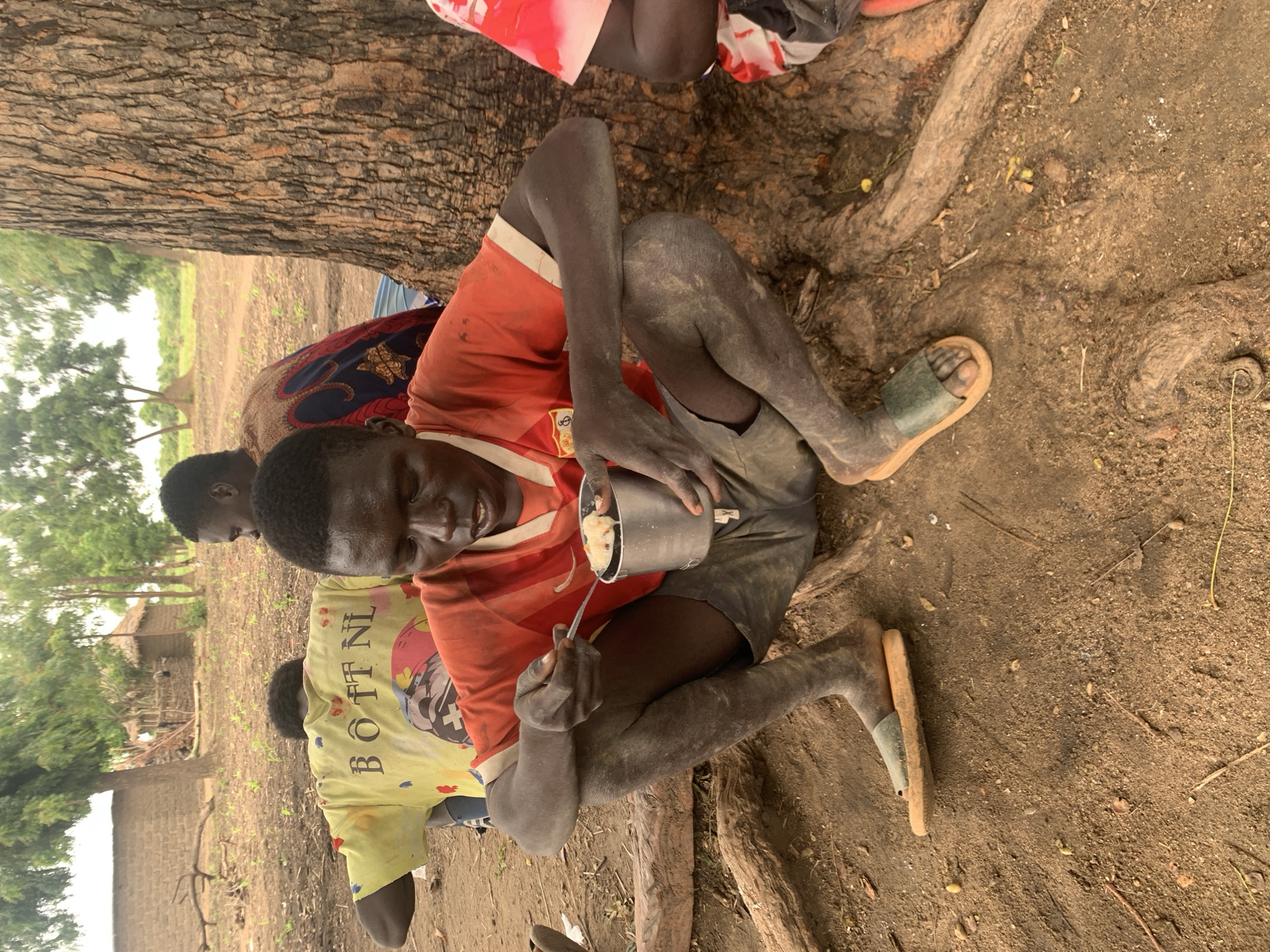 The image shows a young person sitting on the ground, leaning against a tree trunk and eating from a metal bowl with a spoon. The person is wearing an orange t-shirt and grey shorts. Their skin is dark and appears dusty. In the background, other people are partially visible, one wearing a yellow t-shirt with text, and another in a colorful patterned fabric. The ground is sandy and dirt, with exposed tree roots. The setting appears to be outdoors, likely in a village, with some vegetation and rudimentary structures visible in the distance. The lighting suggests it could be daytime, possibly overcast or late afternoon. The overall scene conveys a sense of everyday life, with the focus on the individual eating. The visible text on the yellow t-shirt reads "BOTT NL".