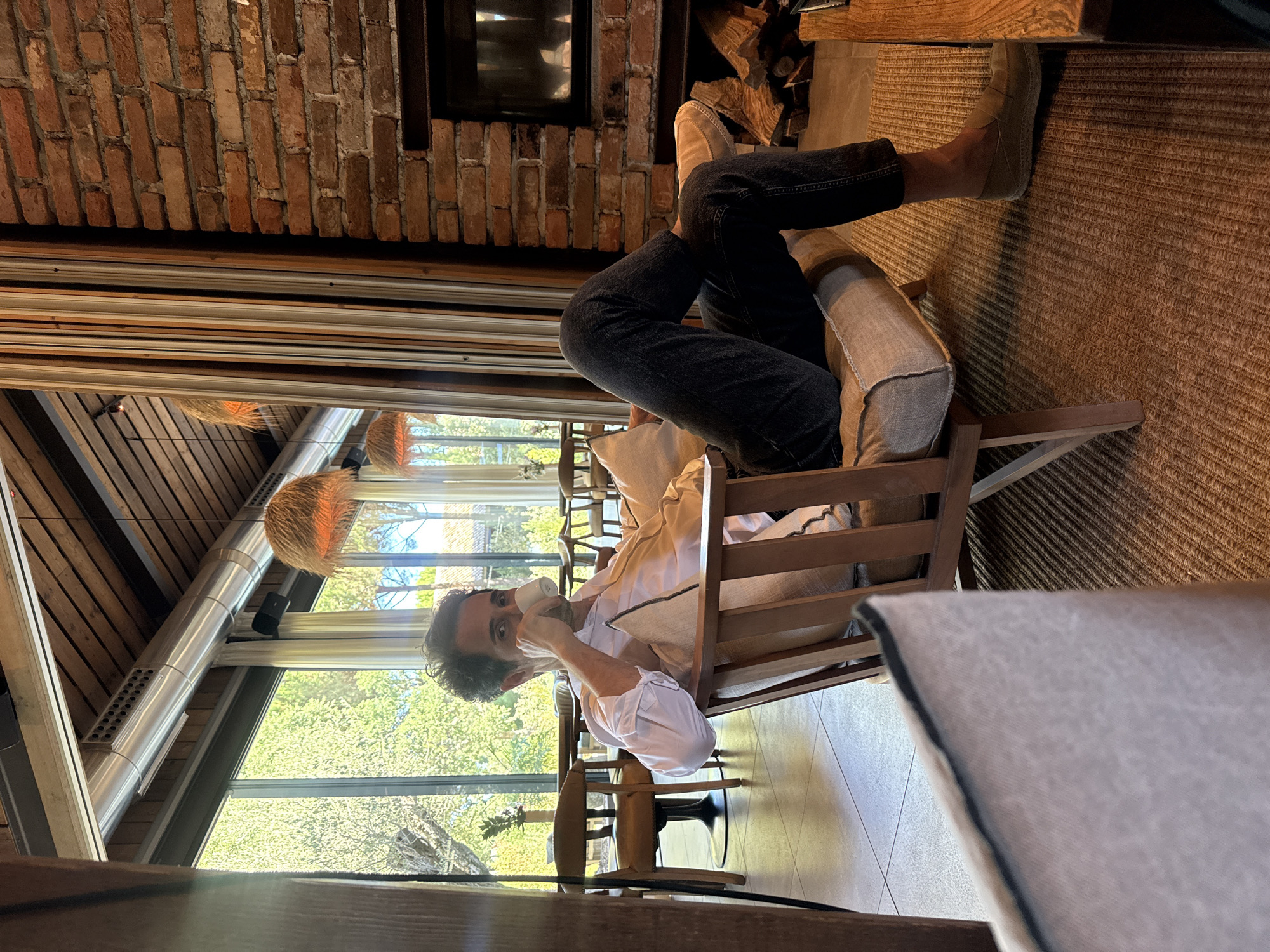 In Grândola, Portugal, a man with curly dark hair is seated in a wooden armchair with a light-colored cushion. He is wearing a light-colored collared shirt with the sleeves rolled up and dark pants. He is holding a white mug to his face with his right hand, appearing to drink from it. The armchair is placed on a textured rug. Behind him, large windows reveal a sunlit outdoor area with trees and greenery. To his right, a brick fireplace is visible, with logs stacked beside it. The room features exposed wooden beams on the ceiling and industrial-style ventilation ducts. Hanging pendant lights with woven shades are also present. The overall ambiance suggests a comfortable, indoor setting with ample natural light.