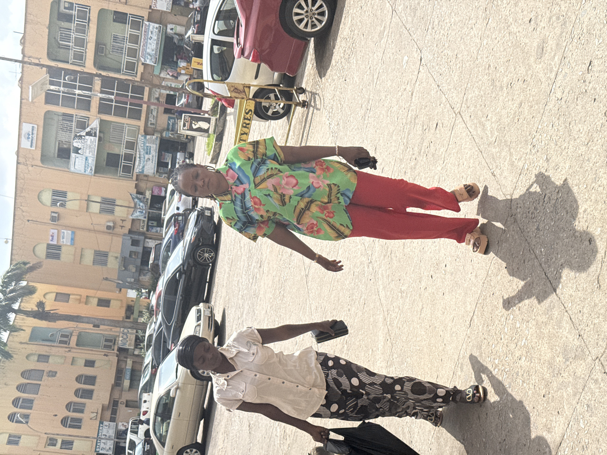 Two women are walking on a concrete sidewalk in Abuja, Nigeria. The first woman wears a green, pink, and yellow floral shirt, red pants, and sandals. She holds a black object in her right hand. The second woman wears a white button-down shirt and black and white polka dot pants. She is carrying a black bag and holds a black object in her right hand. Cars are parked along the side of the road in the background. Buildings line the opposite side of the street, with various advertisements and business signs visible on their facades. The "TYRES" sign is also present. The environment is sunlit, casting shadows on the sidewalk.