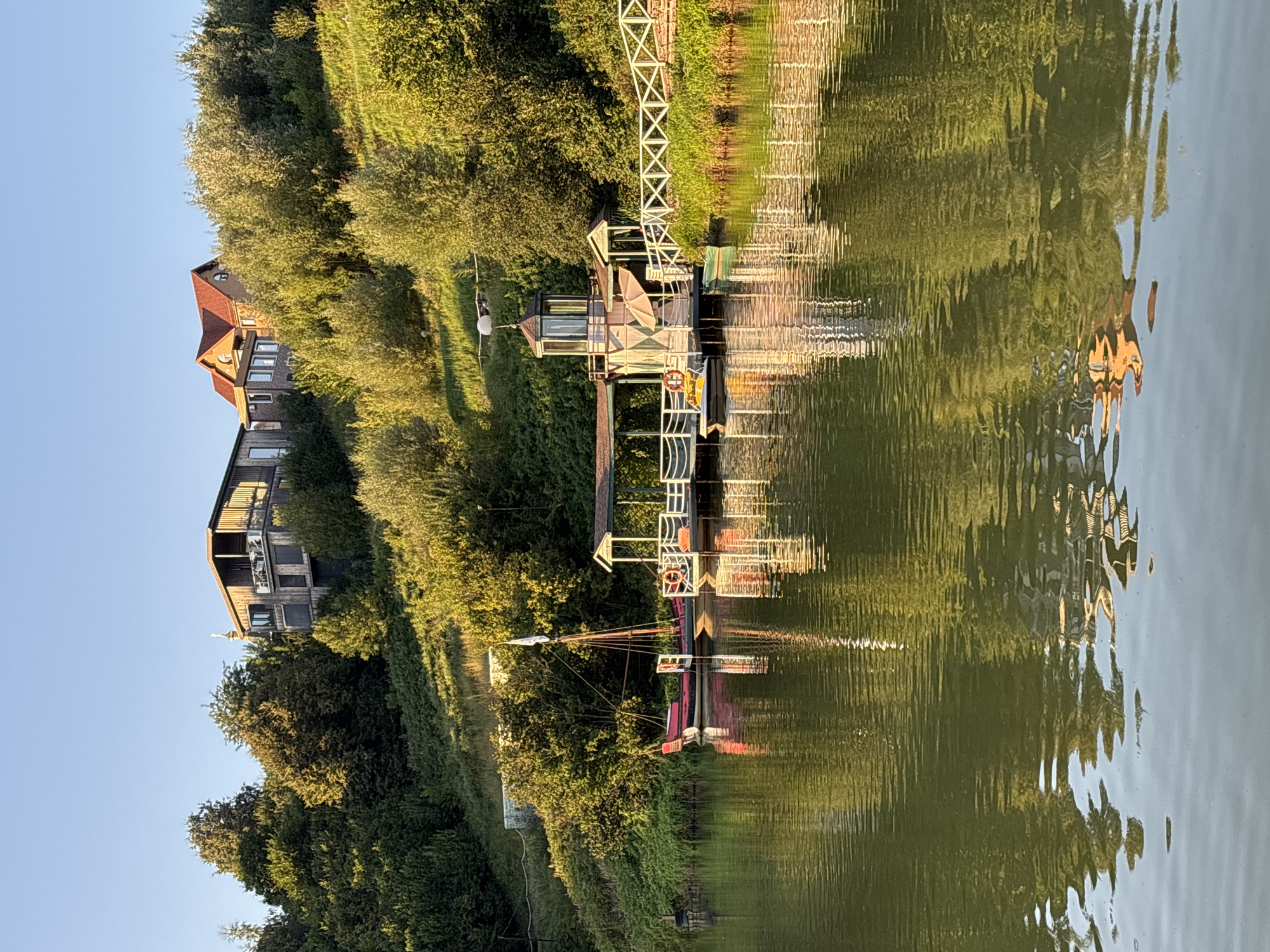 The image depicts a serene waterscape at Romashkovo, Russia, with two modern houses nestled on a lush, green hillside overlooking a calm body of water. The houses feature contemporary architecture with dark wooden exteriors and multiple levels, suggesting luxury residences. In the foreground, a wooden dock extends into the water, adorned with a white railing. Several boats are moored to the dock, including a larger, older-style vessel with a mast and a smaller, yellow paddleboat. The water reflects the surrounding greenery and structures, creating a picturesque and tranquil scene. The sky is a clear, pale blue, indicating fair weather and daylight.
