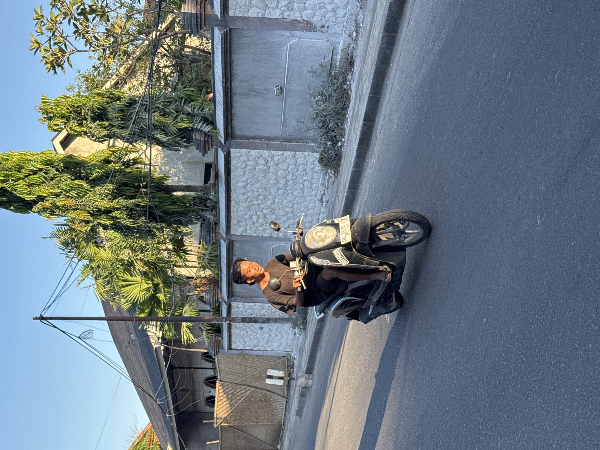 The image shows a man on a black scooter driving on an asphalt road in Kuta, Indonesia. The scene is set during the daytime under a clear blue sky. A variety of trees and buildings are visible in the background along the side of the road. The buildings are constructed with a mix of concrete and stone materials.