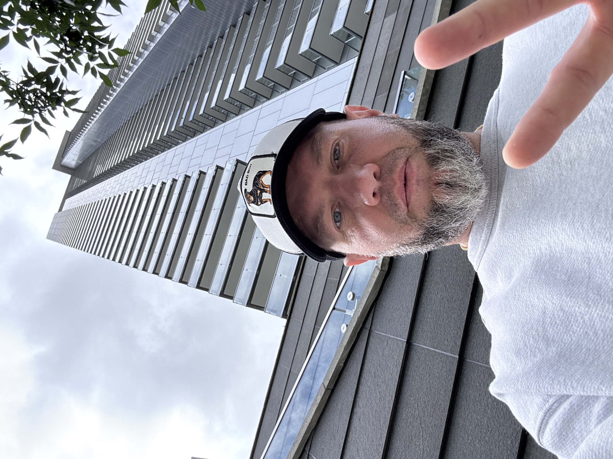 A man with a grey and white beard, wearing a white sweatshirt and a black and white baseball cap with a "BAD BOY" patch, is taking a selfie. He is looking at the camera and making a peace sign with his right hand. Behind him is a modern, multi-story building with balconies and reflective windows, characteristic of a residential high-rise. The building is tall and angular, with horizontal lines formed by the balconies and vertical elements. The sky is overcast. This scene is consistent with the urban architecture found in Burnaby, Canada, a city known for its contemporary developments.