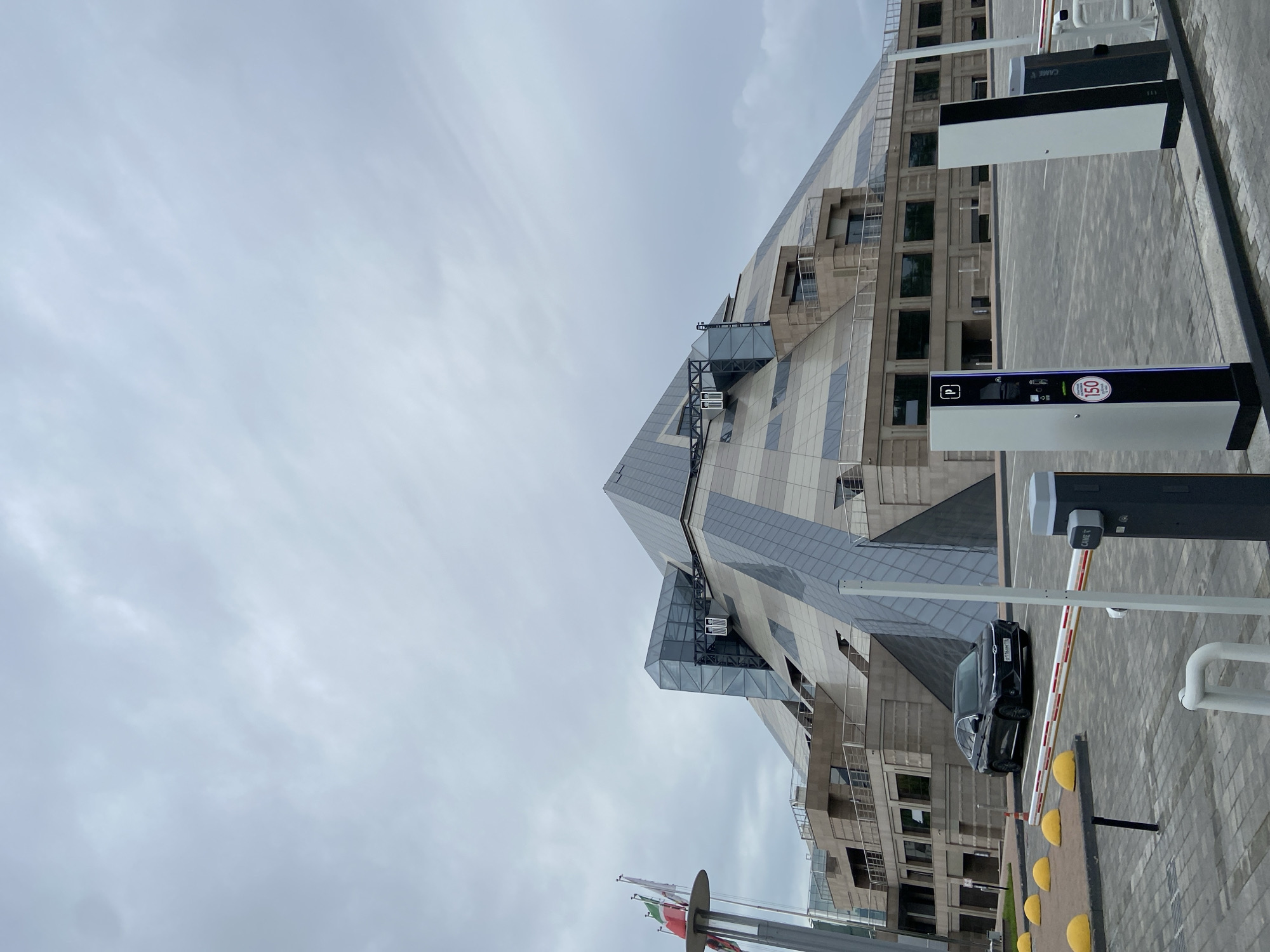 A low-angle, full-shot depicts a modern architectural building in Kazan, Russia under an overcast sky. The structure is distinguished by its angular, geometric design with a mixed facade of glass and light-colored panels.
A parking access control system, featuring automated barriers and payment kiosks, is visible in the foreground. A black sedan is positioned at the barrier, indicating a recent entry or pending exit. Pavement markings and bollards are present near the parking control system.
A flagpole with partially visible flags stands at the lower left corner of the frame.