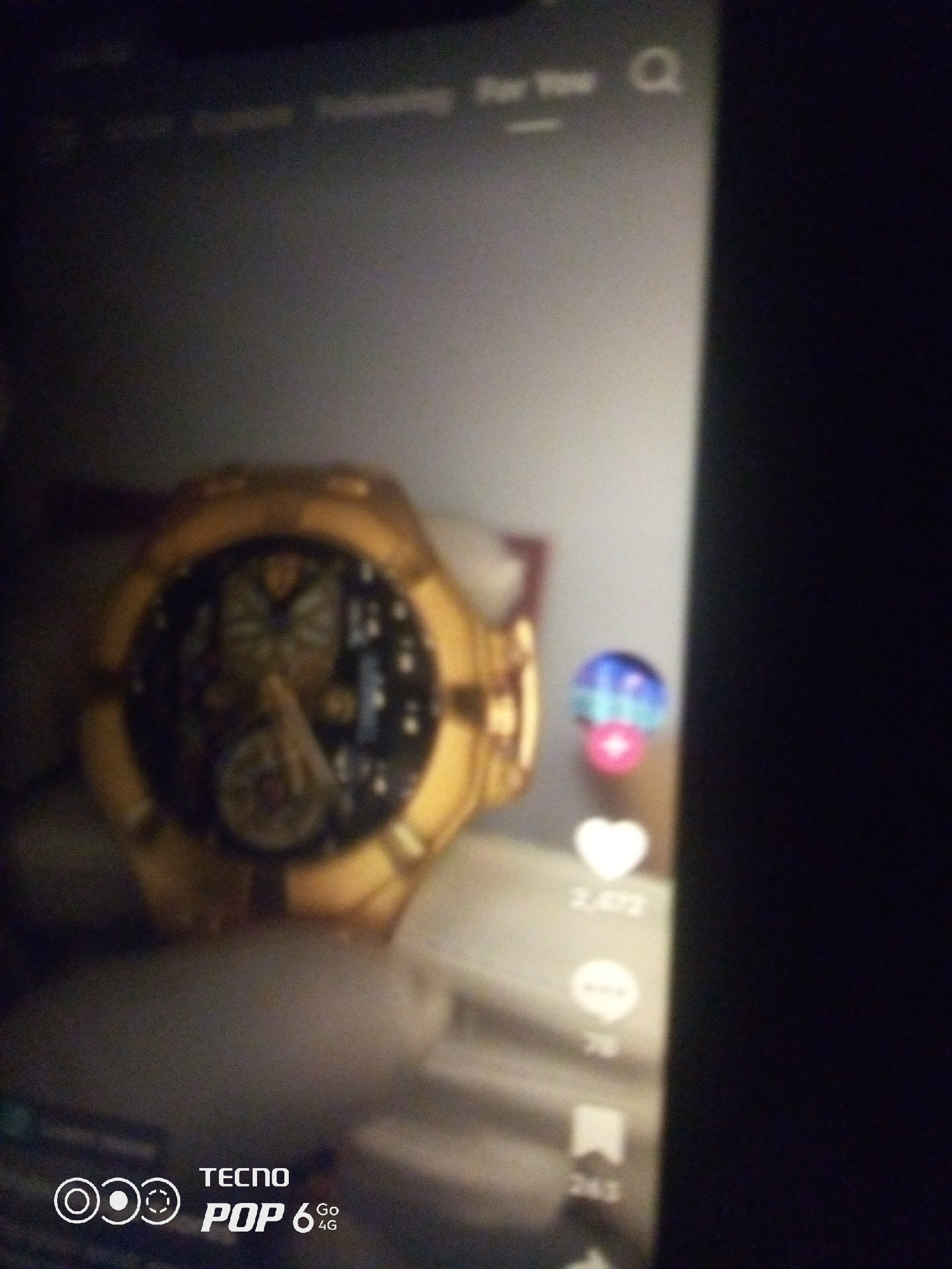 The image shows a close-up of a person's hand holding a golden watch. The watch has a complex dial with various gears and markings. In the background, a screen displays a social media interface with icons for likes, comments, and shares, along with corresponding numbers. The lower portion of the screen shows the "TECNO POP 6 Go 4G" branding. The scene appears to be indoors, with the lighting suggesting it could be a dimly lit room or nighttime. There is no visible information in the image that indicates the location is Howayi, Nigeria.