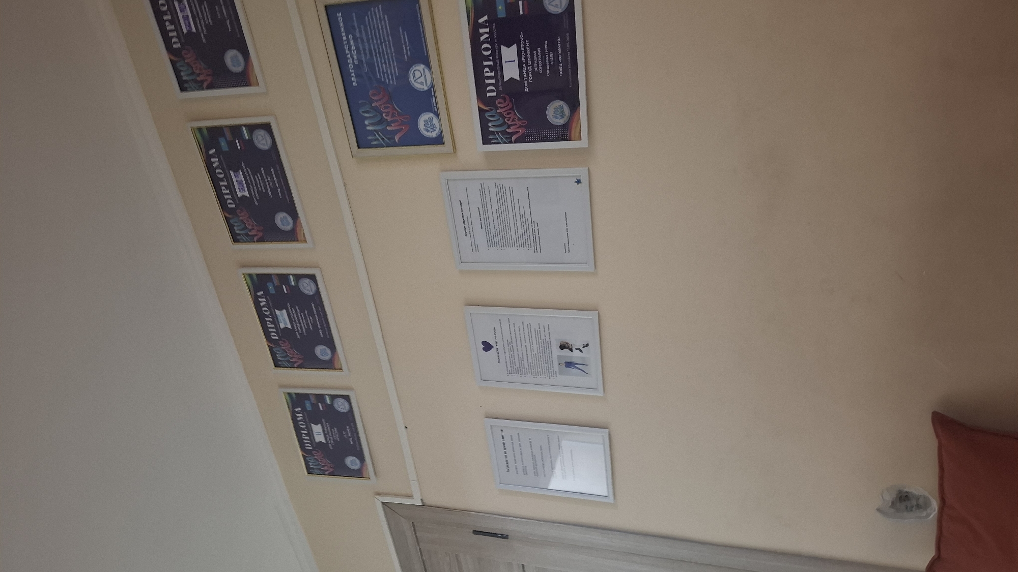 The image displays a wall adorned with several framed documents. At the top, there are four diplomas or certificates, three of which feature prominent text like "DIPLOMA" and "no Vysote." Below these, on the left, is a framed document with text in Russian, seemingly a certificate. Next to it, in the center, is a framed certificate with a purple heart icon at the top, depicting a stylized dancer and text in Russian, including "Уважение родителям!" and "Победителям конкурса." To the right of that is another framed document with Russian text, possibly an award or certificate. Finally, on the bottom right, another framed document with "DIPLOMA" is visible, along with text in Russian including "ДОМ ТАНЦА «FIOLETQVO»," "ГОРОД ШЫМКЕНТ," and "ТАНЕЦ «NO MANTA»." The presence of "ГОРОД ШЫМКЕНТ" (City of Shymkent) in one of the documents places the scene in Shymkent, Kazakhstan. The overall environment appears to be an indoor setting, likely a studio or office, given the displayed certifications.