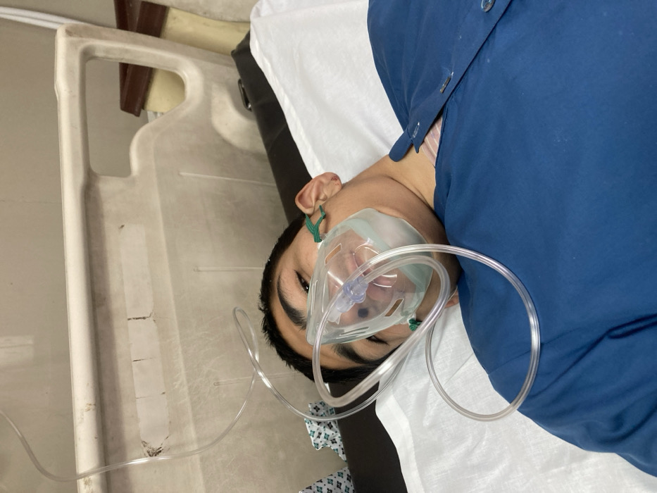 A young male, appearing to be an adolescent or young adult, is depicted lying supine on a bed within a medical facility in Quetta City Tehsil, Pakistan. He has short dark hair and is wearing a clear plastic oxygen mask covering his nose and mouth, secured by green ear loops, with a transparent tube extending upwards and out of frame. He is dressed in a dark blue button-front garment, possibly a patient gown or Kameez, over a lighter-colored, possibly striped, undergarment visible at the neck. He is positioned on a white sheet. Behind him is a light beige/off-white plastic hospital bed rail or headboard exhibiting scuff marks and adhesive residue. A darker brown furniture element, possibly another bed frame, is partially visible in the upper right background. The individual's eyes are open and directed towards the camera.