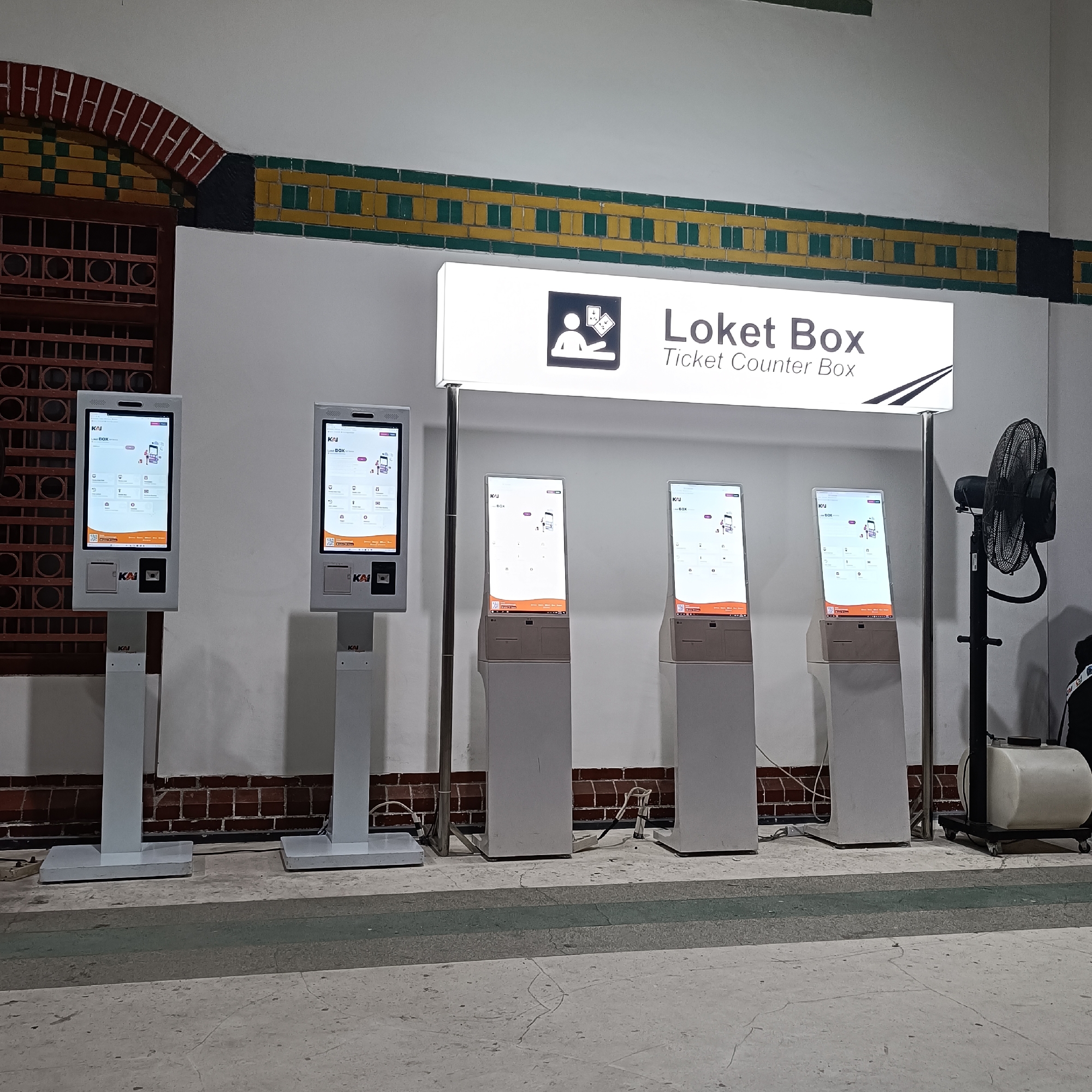 The image depicts an indoor scene at a ticket counter, likely a train station, in Semarang, Indonesia. Four self-service ticket machines, labeled "Loket Box Ticket Counter Box," are arranged in a row. The machines have prominent digital displays showing interface elements, including logos and icons. The background features a white wall with a decorative border of green and yellow tiles, and an arched brick structure on the left. A large industrial fan is visible on the right side of the image. The floor is a patterned concrete surface. There are no people actively interacting with the machines. The overall atmosphere is functional and modern, with a hint of historical architecture in the background.