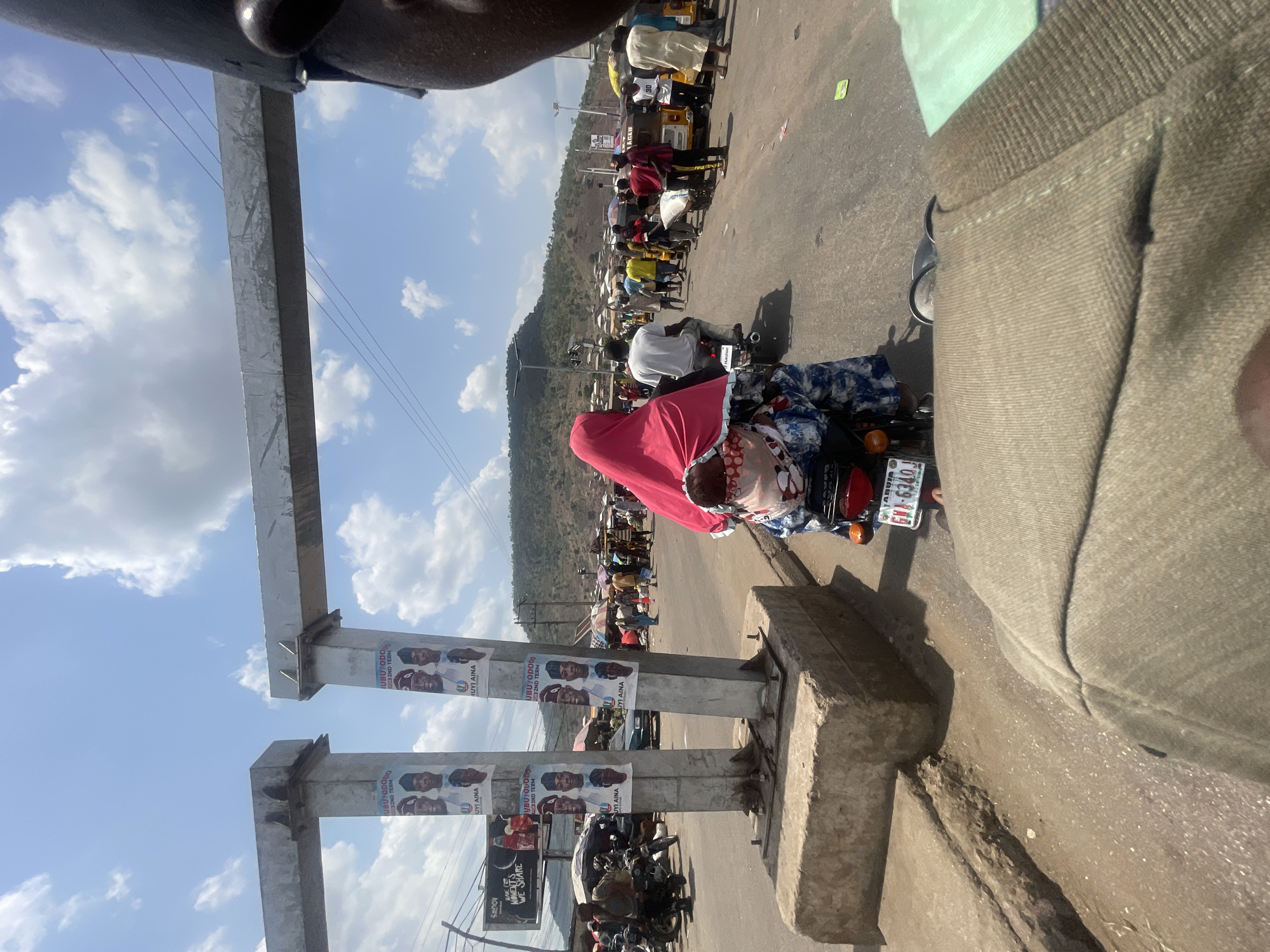 The image shows a street scene in Lokoja, Nigeria. A person on a motorcycle with a red covering over their head is facing away from the camera. The motorcycle's license plate reads "ABUJA GMA-6340J." Political posters for "UBU/ODODO" and "UYI AINA" are affixed to a metal structure. In the background, there is a hill, and the street is populated with people and vehicles, including "Keke Napep" tricycles. The sky is blue with scattered clouds. The photograph appears to be taken from the perspective of another person standing on the side of the road.