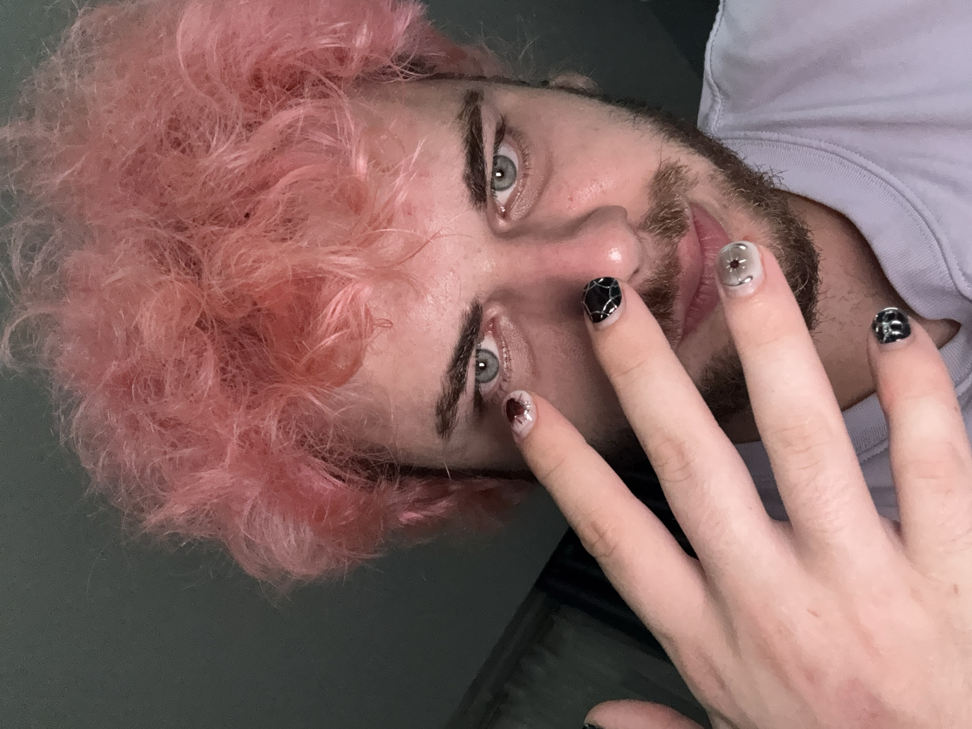 A close-up shot of a person with pink curly hair and a beard. The person is looking at the camera with light-colored eyes. Their right hand is raised, displaying decorated fingernails with dark designs. The background appears to be a plain wall. Location information is not discernible from the image.