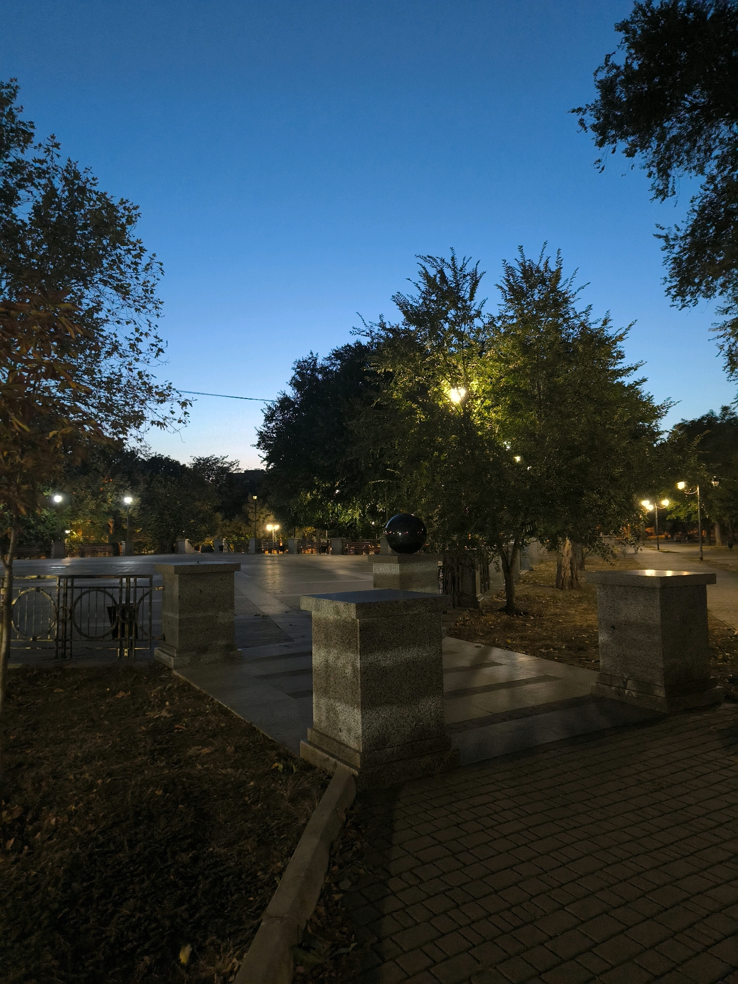 The image depicts a public park area at dusk or early dawn, with the sky a deep, clear blue. The park is illuminated by street lamps, casting a warm glow on the trees and walkways. Several trees with dense foliage are prominent, some with lights embedded within their branches. A large, dark, spherical sculpture is visible on a pedestal in the foreground, partially obscured by a tree. Paved pathways and stone structures, possibly benches or decorative elements, are scattered throughout the scene. The ground is covered with dry leaves in some areas, suggesting an autumn season. In the background, more trees and lighting fixtures are visible, creating a serene, ambient atmosphere. The location is identified as a park in the city of Mykolaiv, Ukraine. There are no people or animals present in the image, and no overt actions or interactions are taking place. The overall impression is one of a quiet, well-maintained urban green space.