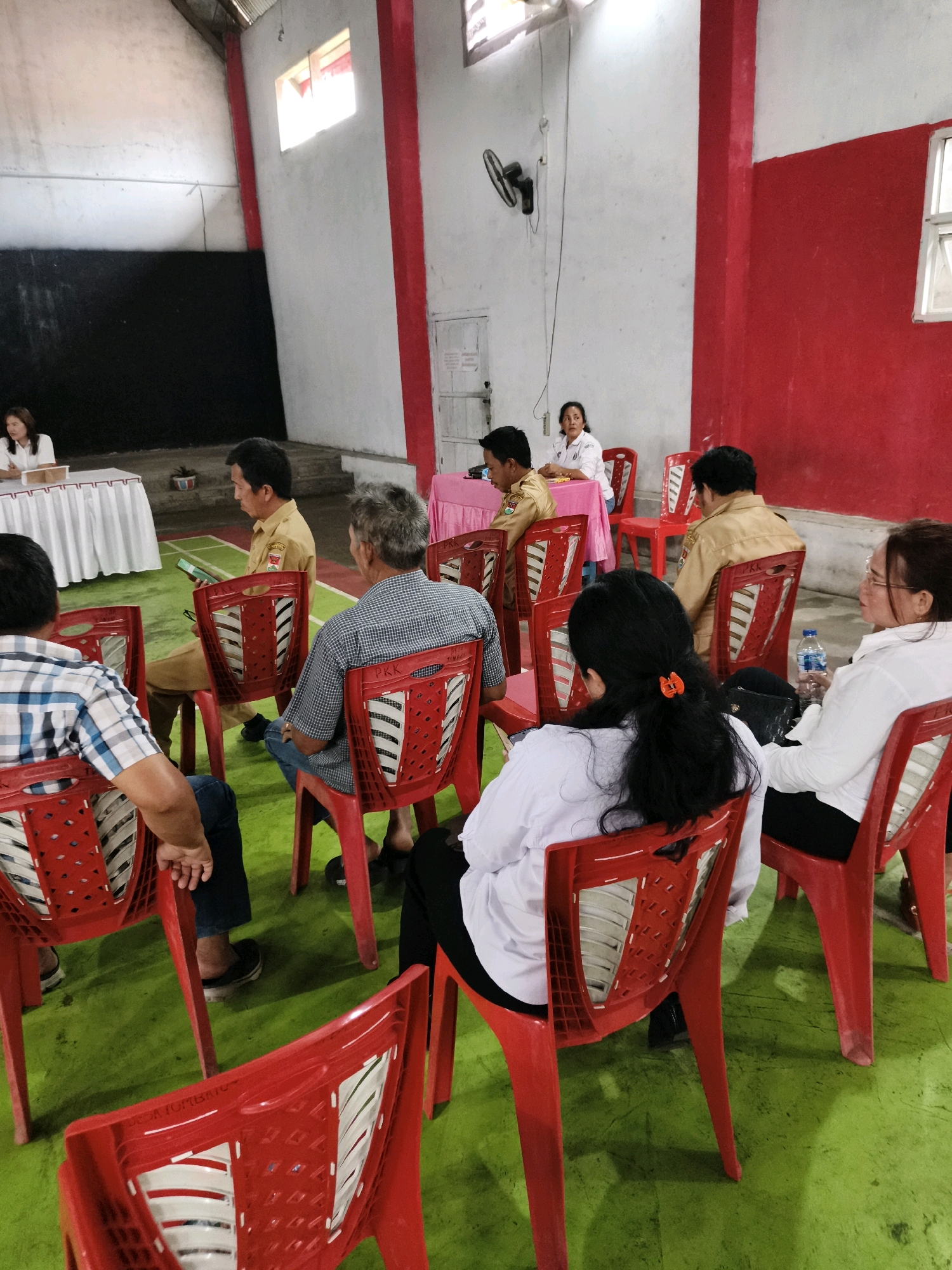 In Tombatu Satu, Indonesia, a group of people are seated in red plastic chairs on a green floor in a large indoor space. The walls are predominantly white with red accents. In the background, on a raised platform, two women are seated at a table covered with a white cloth, facing a few other individuals. One woman wears a white blouse, and the other, a white polo shirt with a logo. To the right, a man in a beige uniform is seated, holding a green tablet. Several other individuals, mostly men, are seated in red chairs facing away from the camera, some wearing white polo shirts. A woman in a white shirt and black pants is visible on the far right. Another woman with long dark hair is seated in the foreground, also facing away from the camera. The room appears to be a community hall or meeting space.
