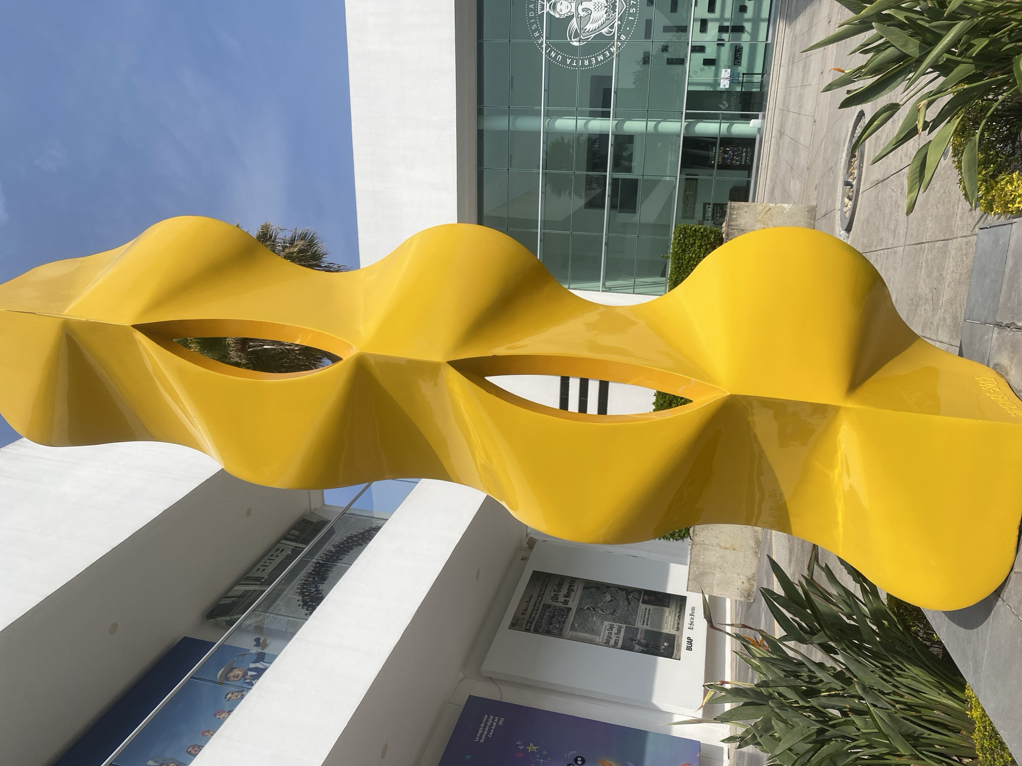 A brightly lit, outdoor, vertically-oriented shot features a vibrant yellow, wavy, abstract sculpture dominating the foreground and mid-ground. The sculpture, which has several open, oval-shaped cutouts, appears smooth and reflective.
In the background, a modern building with large, reflective glass windows and white facade is visible. The windows reflect the bright blue sky and elements of the surroundings. A circular university seal with text, partly readable as "BENEMERITA UNIVERSIDAD," is visible on the upper section of the building's glass facade.
The scene is illuminated by bright daylight, suggesting a sunny day with a clear blue sky, parts of which are visible above the sculpture and reflected in the building. Lush green plants, including palm fronds and other leafy vegetation, are positioned around the sculpture and near the building. Paved ground covers the area around the base of the building and sculpture.
Visible text includes:
* On the building's reflection, part of a university seal: "BENEMERITA UNIVERSIDAD".
* On white panels near the building's base: "La magia de crear ilustración digital Coco Audi Art 2023", "Un Golazo de Negretel", "Bélgica Dejó Fuera a Rusia!", and "BUAP El Sol de Purita".
* On the sculpture itself, barely legible: "2000-2002".
No specific activity or people are depicted in the static scene, which highlights public art within what appears to be a university campus setting.