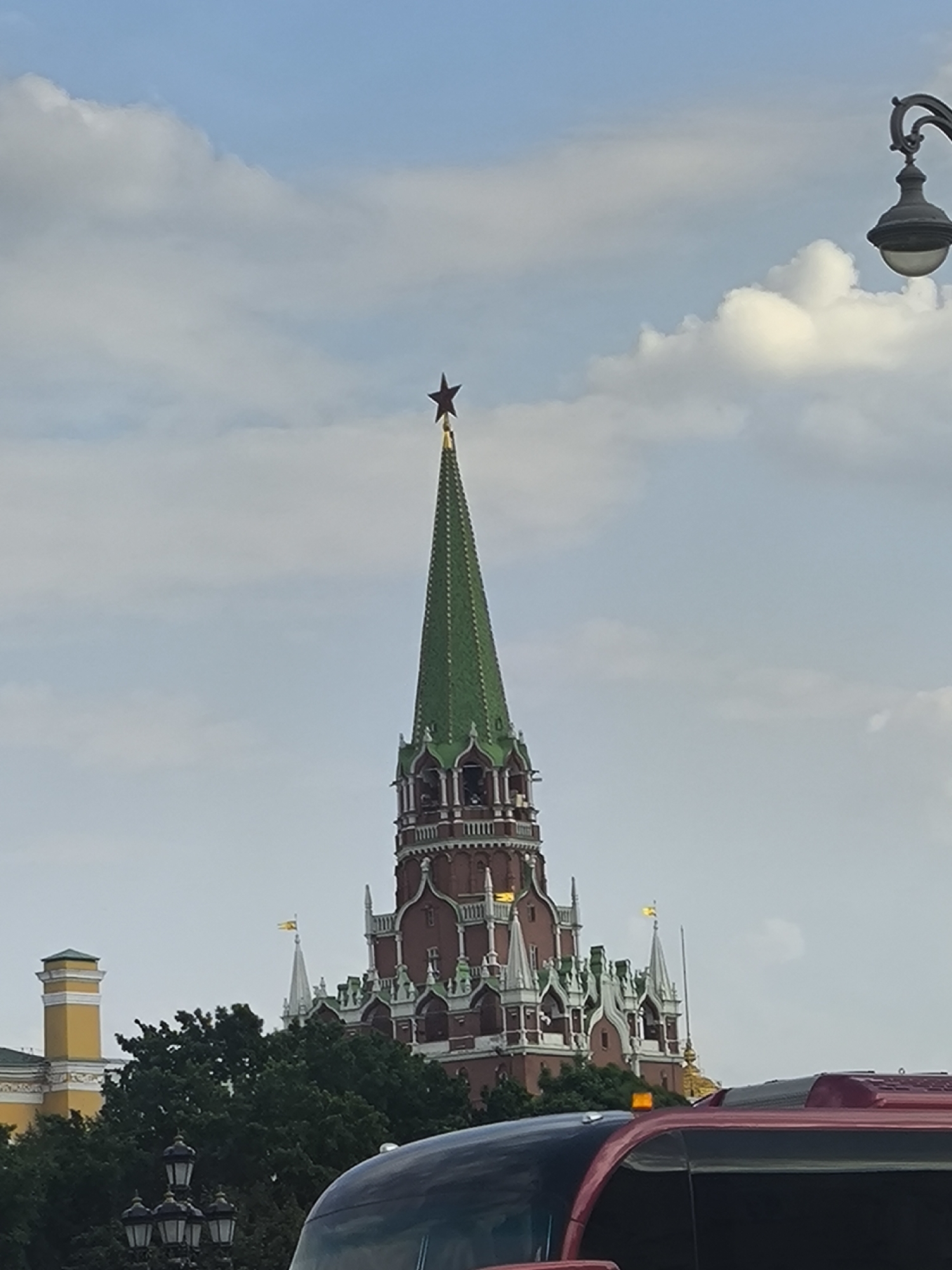 The image depicts a tower in Moscow, Russia, likely part of the Kremlin. The tower is tall and slender, with a green roof and a red star at the top. It is ornately decorated with white and green details. In the foreground, there are trees, a lamppost, and a portion of a red and black vehicle, likely a bus or large car. The sky is visible in the background, with light blue and white clouds.