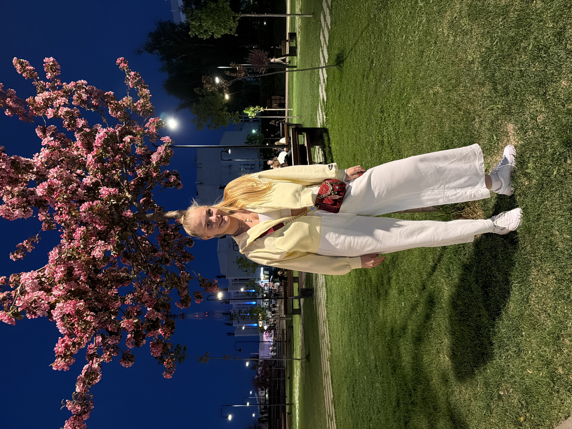 A woman stands in a park in Izhevsk, Russia, at night. The park has benches, trees, and grass. The sky is dark blue. Street lights illuminate the scene. A large tree with pink blossoms is on the left side of the frame. The woman is wearing white pants, a white top, and a pale yellow jacket. She has a small red purse on her shoulder. The woman is smiling and looking at the camera.