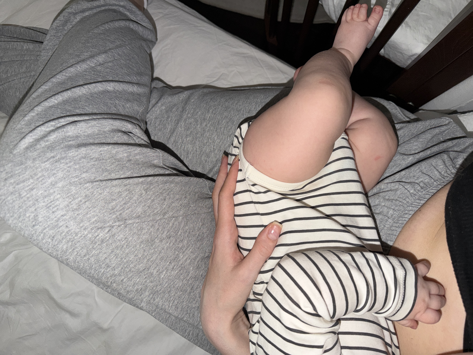 An intimate, high-angle shot captures an adult holding a baby in what appears to be an indoor setting, likely a bedroom. The adult's lower body, clad in grey marl sweatpants, occupies the left side of the frame. Their hand is gently placed on the baby's back/side, providing comfort.
The baby, wearing a white long-sleeved onesie with thin black horizontal stripes, lies on the adult's lap or chest. Its small, chubby legs and feet are prominent, with one foot extending upwards towards the right, close to a dark wooden structure, possibly a crib rail or headboard. A faint, small red mark is visible on one of the baby's thighs.
The background shows rumpled white bedding, reinforcing the bedroom environment. The lighting is soft and ambient, suggesting a cozy, possibly low-light time of day, such as evening or night. The overall scene conveys a feeling of warmth, affection, and peaceful parental care. There is no visible text in the image. This media file was taken in Khimki, Russia.