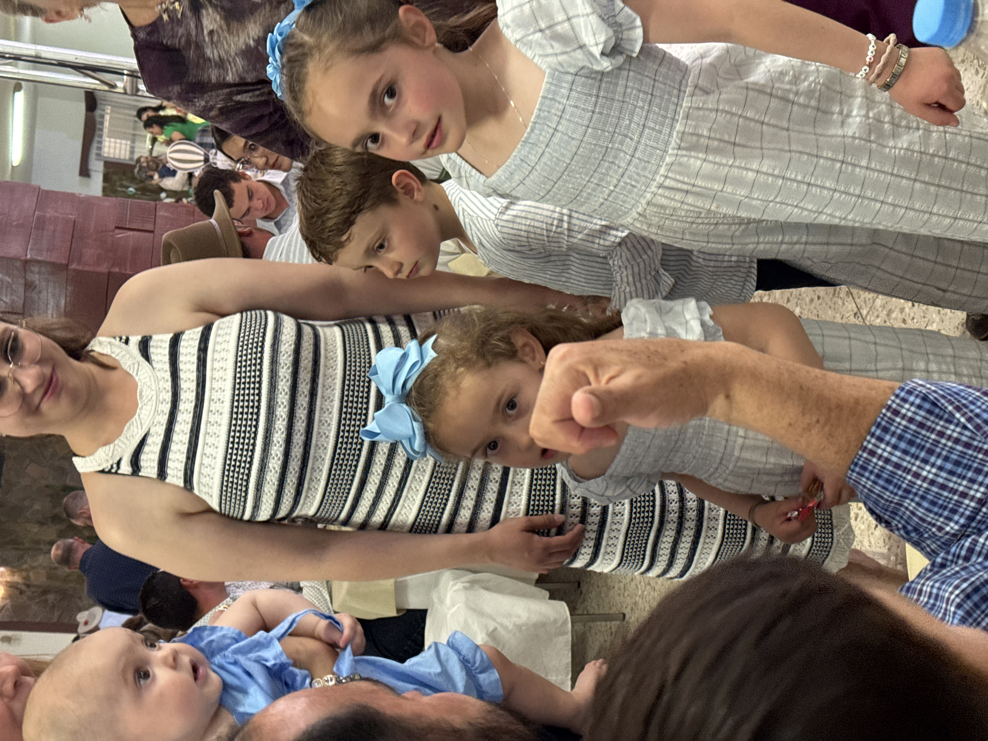 The image captures an indoor scene with multiple individuals. The foreground features a baby wearing blue, held by an adult, and a woman in a black and white striped dress. Three children are also visible: two girls, one with a blue bow in her hair, and a boy, all in striped or patterned clothing. One of the girls has bracelets on her right wrist. An adult is also partially visible, wearing a blue plaid shirt.
Background details include other people, some wearing hats, and what appear to be rows of objects, possibly boxes or containers, stacked along a wall. Fluorescent lighting is visible on the ceiling. The image appears to have been taken at an event. Without additional details, specific events or locations in Chipilo are indeterminable.