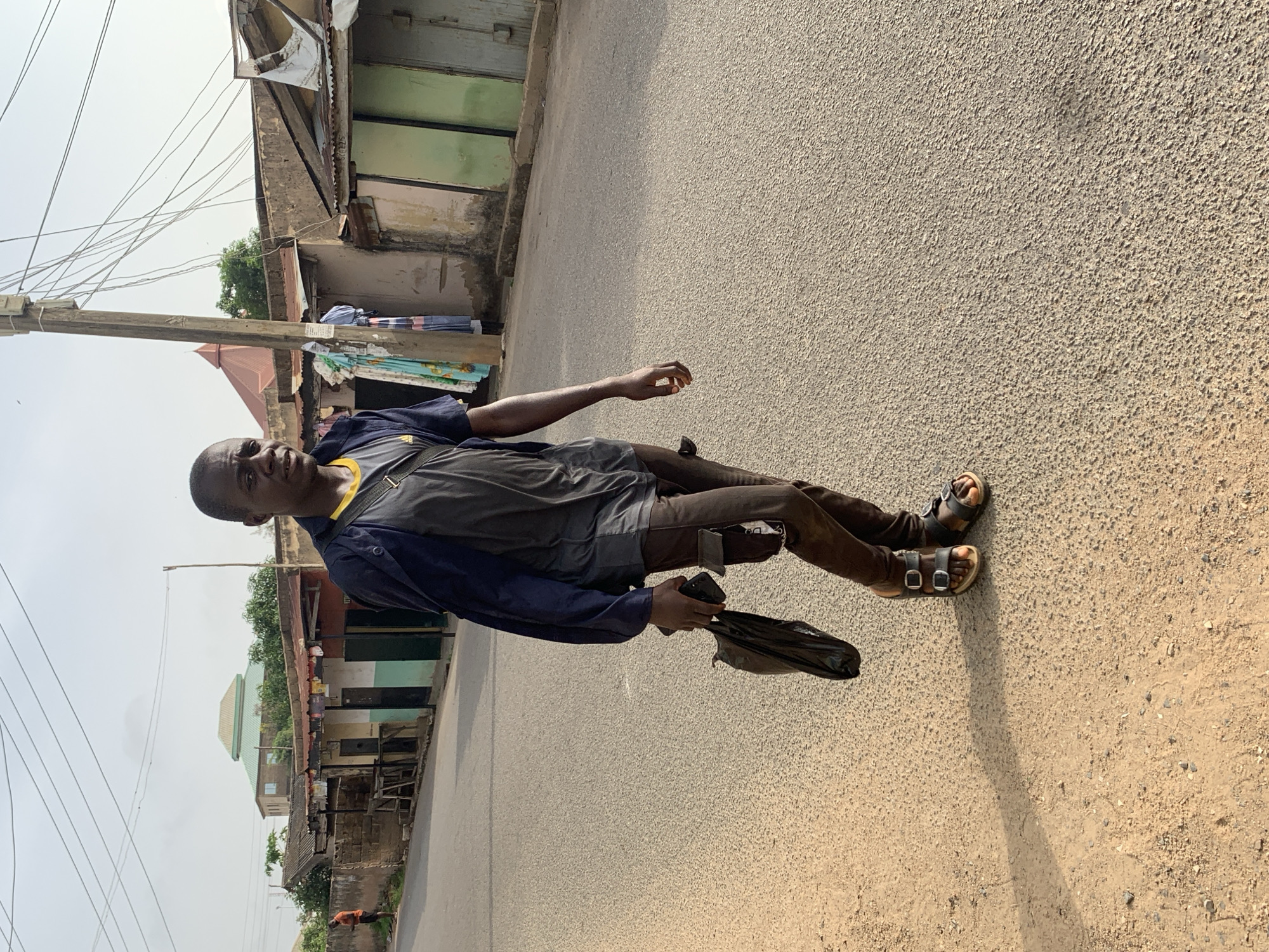 A man stands on a paved street in Jos, Nigeria. He is wearing sandals, brown pants, a gray shirt, and a blue jacket. He holds a black plastic bag. The background shows buildings with corrugated iron roofs and power lines overhead. The sky is overcast.