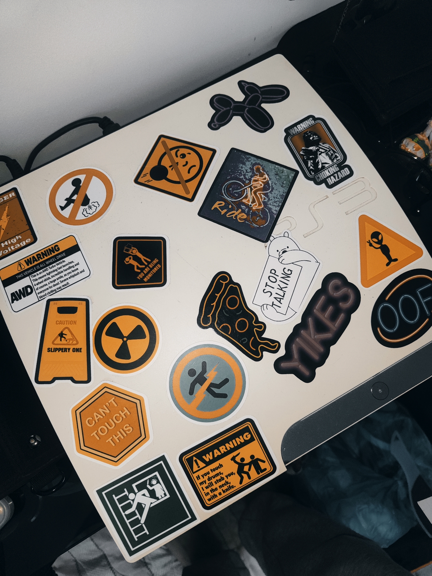 The image shows a close-up, high-angle view of a laptop covered in various stickers. The laptop is open and resting on a dark surface, possibly a desk or a bag. The background is mostly out of focus, with some blurry dark objects and a white wall visible. The stickers are colorful and diverse, featuring a range of humorous and warning symbols.
Notable stickers include:
- A balloon dog.
- A warning sign with a figure being electrocuted.
- A sign depicting a figure being sprayed with gas, with "NO" over it.
- A sticker of Darth Vader with "WARNING CHOKING HAZARD" text.
- A sticker of a person riding a bicycle with the word "Ride".
- A sticker of a slice of pizza.
- A sticker of a white bear holding a sign that says "STOP TALKING".
- A yellow triangular sign with an alien figure.
- A neon sign that says "OOF".
- A dark sticker with the word "YIKES".
- A yellow hexagonal sign with "CAN'T TOUCH THIS" text.
- A caution sign with a slippery floor symbol and "SLIPPERY ONE" text.
- A yellow sign with a radioactive symbol.
- A large white and yellow sticker with "WARNING THIS IS AN AWD Turbo Vehicle. This means lightning fast launches. 4 wheel slides. Incredible handling and of course, a huge smile, as you leave everyone for dead. Severe whiplash and blood clotting may result." and the text "AWD".
- A square sticker with two figures and the text "YOU ARE BEING MONITORED".
- A black and white sticker depicting a figure climbing a ladder towards a window where another figure is visible, with what appears to be warning text in the bottom right corner that is partially obscured.
The image is taken indoors, likely in a personal space. There is no indication of a specific location in Tashkent, Uzbekistan. The scene is static and depicts inanimate objects.