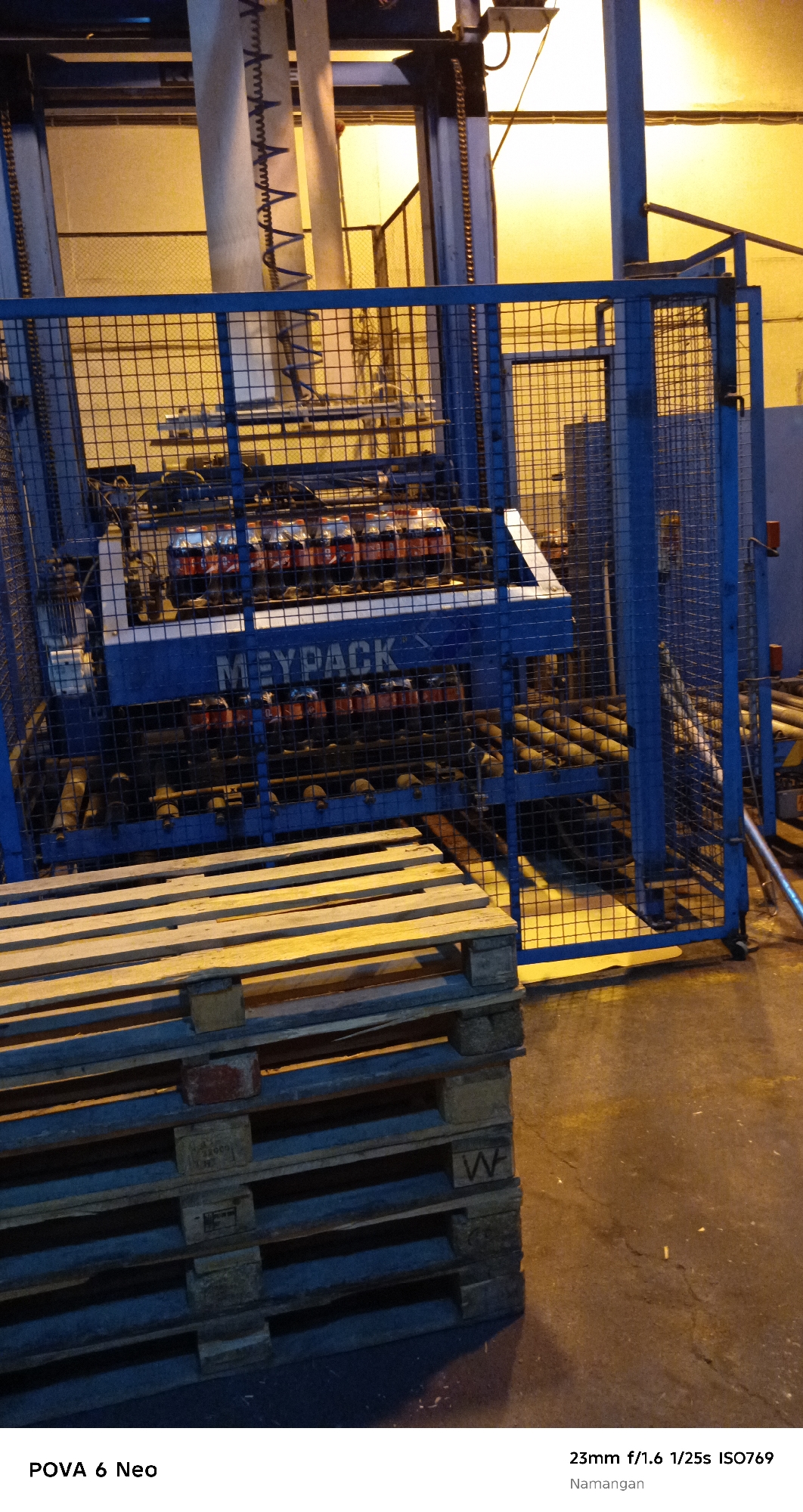 A pallet of wooden planks is in the foreground, to the left. Behind it, a blue metal cage encloses a beverage bottling or packaging machine, identified by the "MEYPACK" logo. Rows of bottles are visible within the machine, illuminated by ambient yellow light. The machine is situated in what appears to be a factory or warehouse setting, evidenced by industrial equipment and the general ambiance. The ground is a concrete floor. The photograph was taken in Namangan City, Uzbekistan, with a camera setting of 23mm f/1.6, 1/25s, and ISO 769.