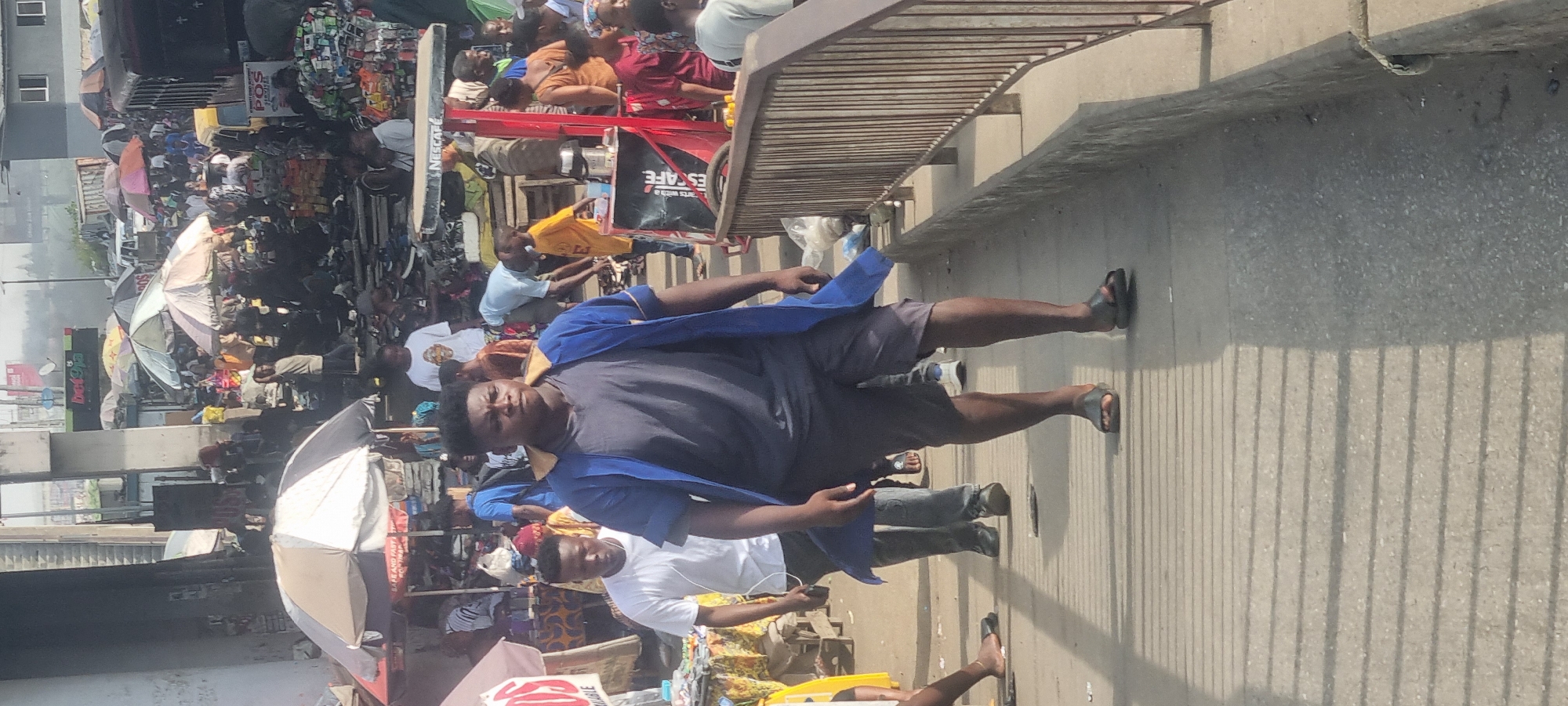 The image shows a busy scene on a footbridge in Ikeja, Nigeria. A man in a dark t-shirt, grey shorts, and a blue open shirt is standing on the bridge, facing the camera. A younger man in a white t-shirt is standing slightly behind and to the left of him. The footbridge has a grey surface and metal railings. Below the bridge is a crowded market area with numerous people, stalls, umbrellas, and goods for sale. There are signs and posters visible in the market. The sky is bright, suggesting daylight.