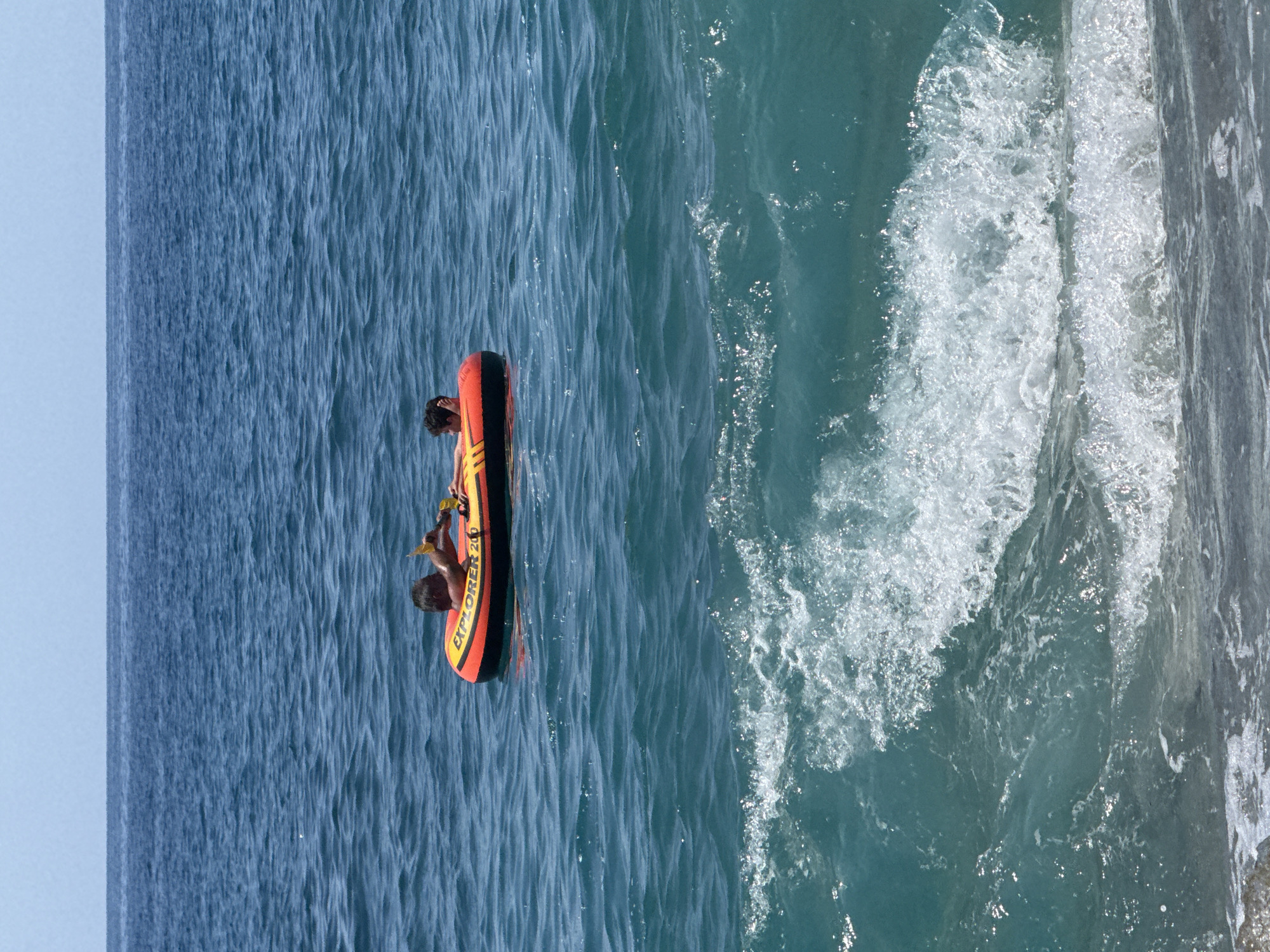 Two children, one on each side, sit in a bright orange and yellow inflatable tube with "EXPLORER 200" written on it. They are in the middle of a vast expanse of deep blue water under a clear blue sky. The water is calm with a slight ripple. In the foreground, a white wave is crashing, obscuring the sandy shore below. The scene suggests a recreational activity at a beach, likely in Alanya, Turkey, given the clear, warm weather and typical beach activities.