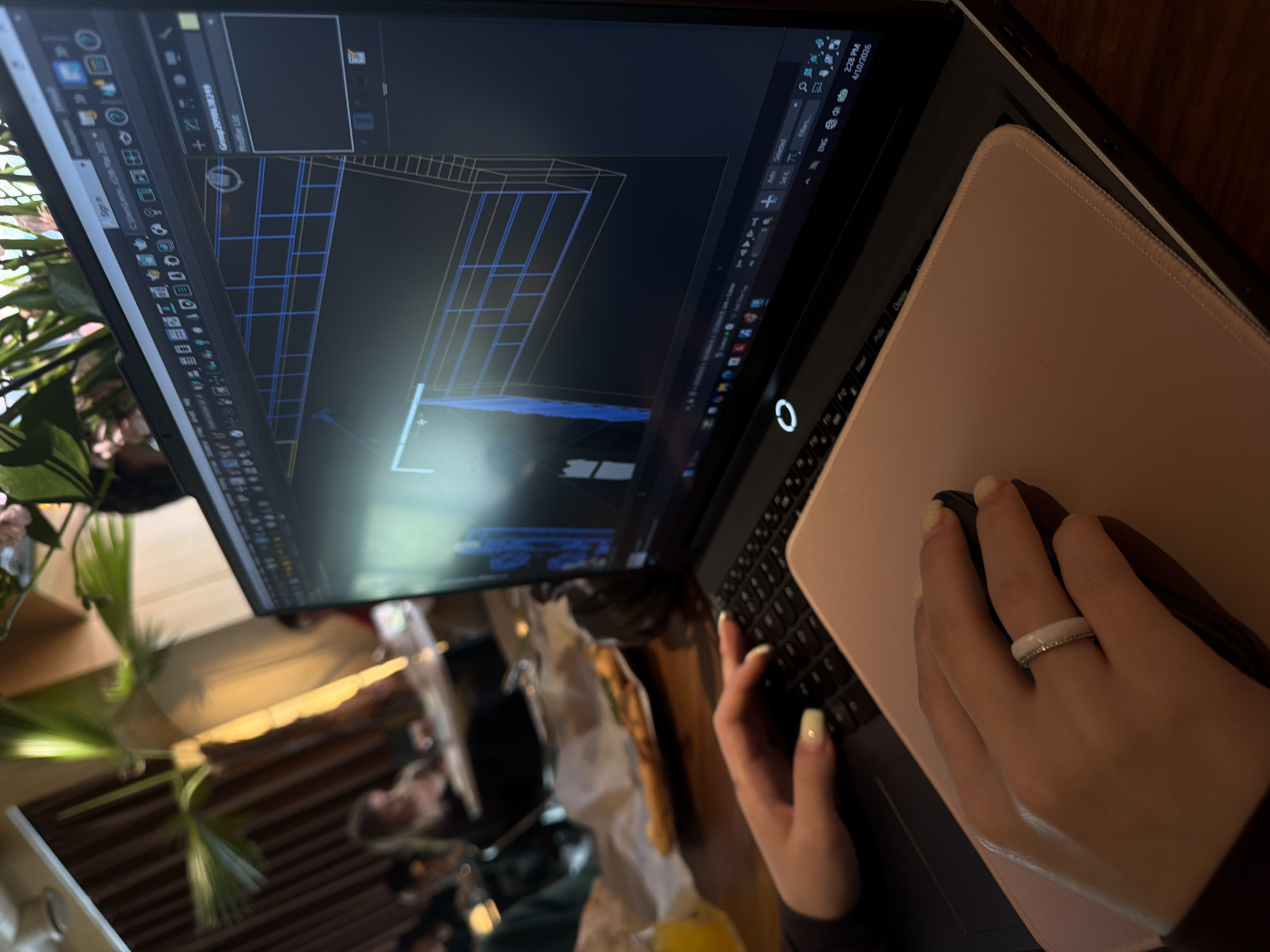 The image shows a person using a laptop computer. The laptop is open and displaying a 3D modeling program. The person's hands are visible, with one hand using a mouse on a pink mousepad. The person is wearing a white ring on their right hand. In the background, there is a plant and what appears to be a cafe or restaurant setting, with other people seated and possibly eating. The timestamp on the laptop screen shows the date as April 10, 2026, and the time as 2:28 PM. The city of Yerevan, Armenia is not explicitly mentioned or identifiable from the provided visual information.