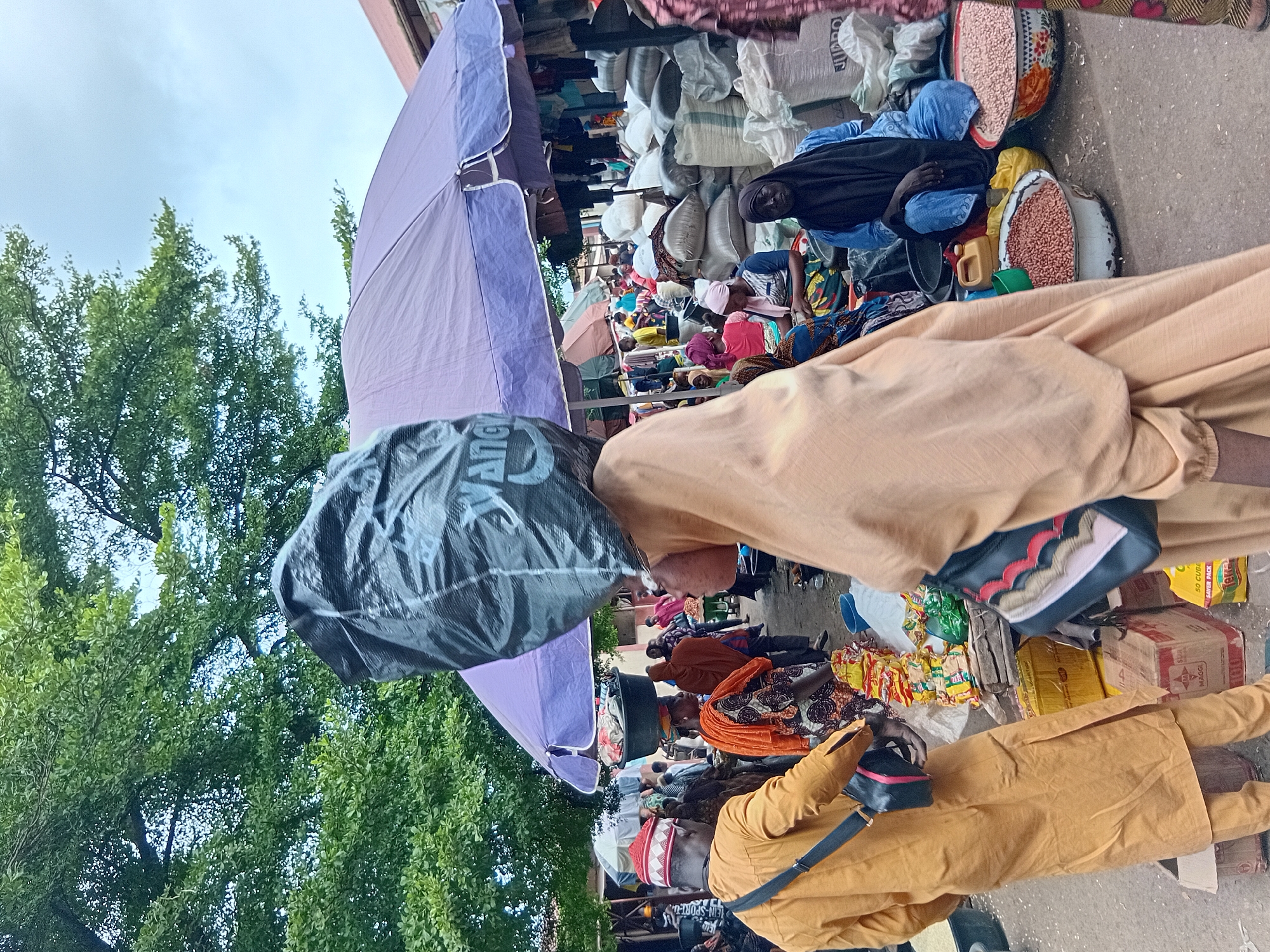 This is a medium shot, taken outdoors, at eye-level, in a busy market in Lokoja, Nigeria. The scene is filled with people and goods. In the foreground, a person wearing a tan-colored robe and a headscarf is walking away from the camera, carrying a large black trash bag on their head. Another person in a bright yellow robe and a red and white patterned cap is walking in the same direction, with a black bag slung over their shoulder.
The background is a bustling marketplace with numerous stalls and people. Large purple and green umbrellas provide shade. Various goods are on display, including sacks of grains or other products, bowls of red beans, and packaged food items like Maggi seasoning. Several individuals are visible, some sitting and some standing, engaged in market activities. The ground is dusty and appears to be dirt. Trees with green foliage are visible in the upper portion of the image, suggesting an outdoor environment. The overall impression is one of a lively, active marketplace.