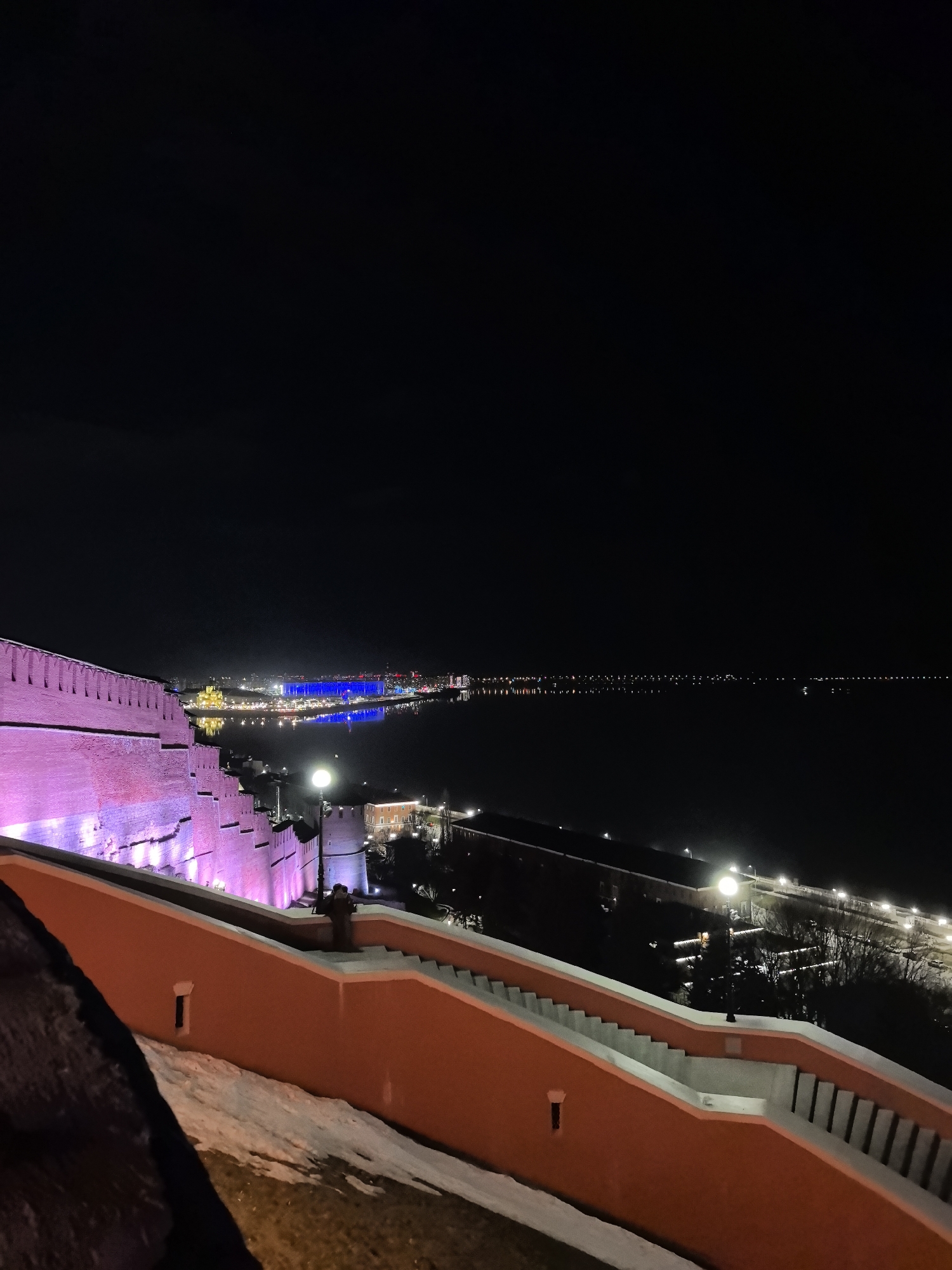 The image captures a night scene in Nizhny Novgorod, Russia. A portion of the Nizhny Novgorod Kremlin wall is illuminated with pink light on the left side of the frame. Adjacent to it, stairs with white edges descend from the wall, colored in a light-orange hue. Snow patches are visible beside the stairs.
In the background, a body of water extends horizontally across the frame, reflecting the lights of the cityscape on the opposite shore. The city lights include a striking illuminated blue building. Along the near shore, streetlights are spaced out along a road.
The sky above is dark.