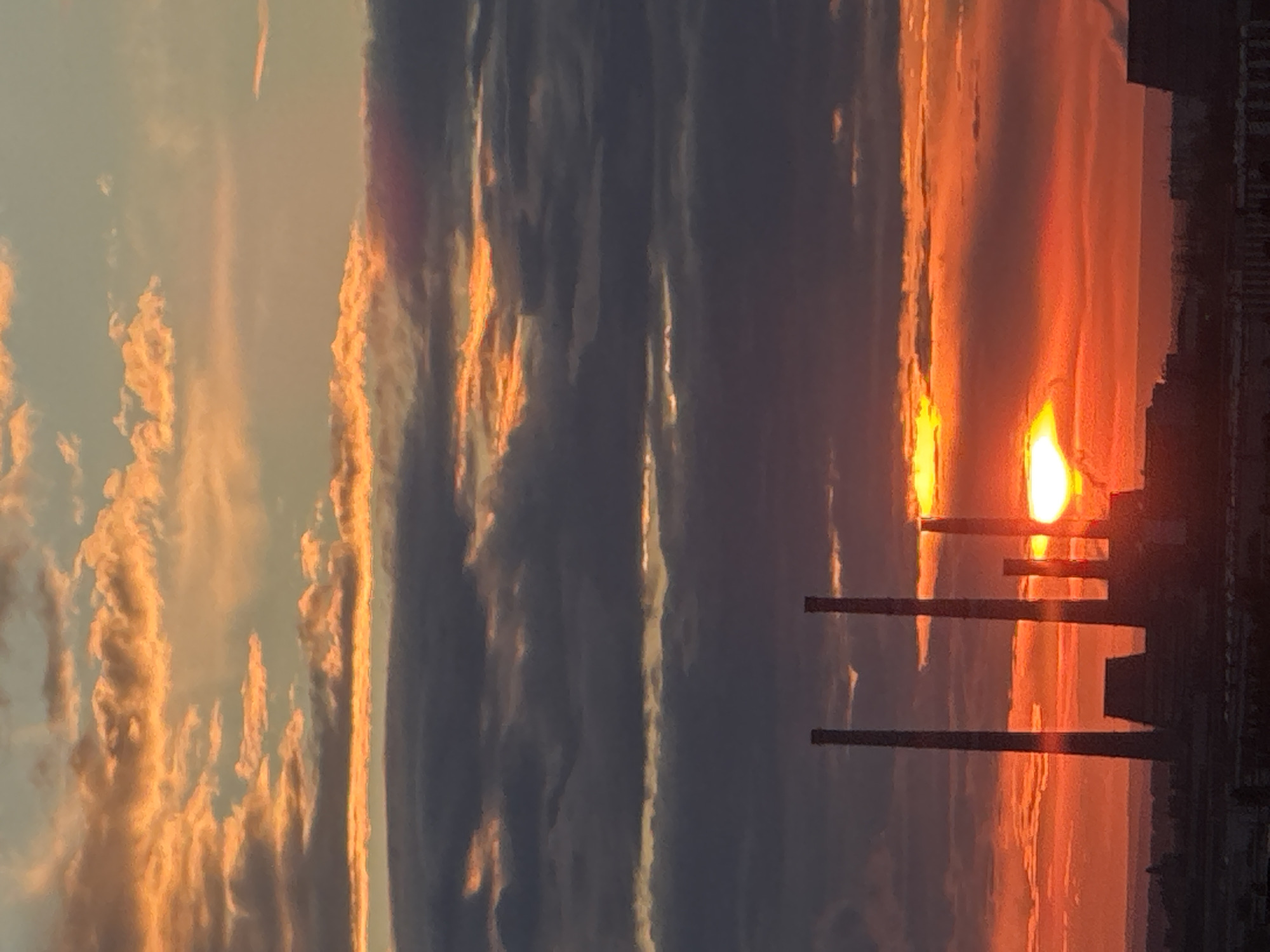 A dramatic sunset casts a fiery orange glow across the sky, illuminating a cluster of industrial smokestacks that stand silhouetted against the vibrant clouds. The clouds themselves are a mixture of deep grays and bright oranges, with streaks of sunlight breaking through the darker masses. The smokestacks, which appear to be part of a power plant or factory, are tall and cylindrical, with one of them emitting a faint plume of smoke. In the foreground, the rooftops of a city are visible, suggesting an urban environment. The overall impression is one of industrial might meeting the power of nature in a breathtaking display of light and shadow.
