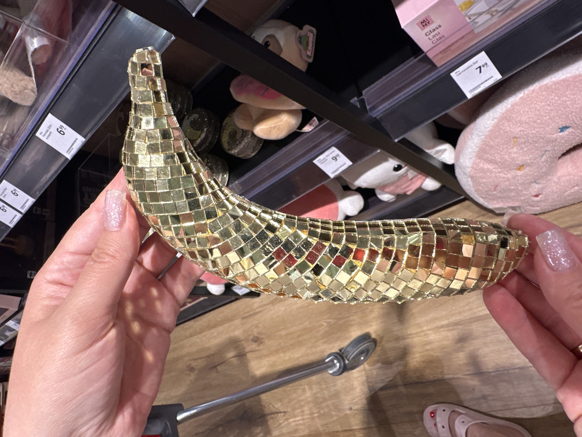A person is holding a gold, sequined banana-shaped object in their hands. The object is decorated with small, square, mirrored tiles that reflect light. The background shows a store aisle with shelves stocked with various items, including plush toys and other decorative objects. A price tag with the number "6.99" is visible on a shelf in the upper left corner of the image. Another price tag with "7.99" is visible on a shelf in the upper right. A shopping cart handle is visible in the lower portion of the image. The lighting suggests an indoor setting, likely a retail store. The person's hands are manicured with glittery nail polish, and they are wearing a ring on their right hand. The person is also wearing a pale pink shoe. The image is taken from a close-up perspective, focusing on the banana object and the person's hands.