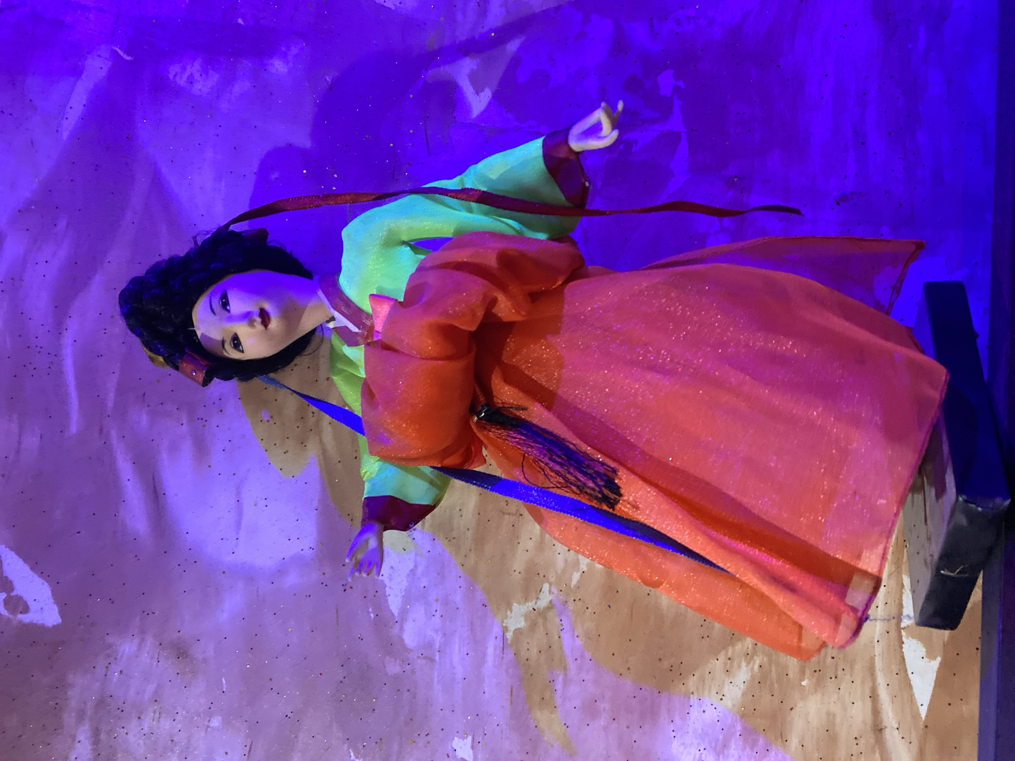 This image features a doll, posed vertically, depicting a female figure with pale skin, dark hair styled in an updo, and distinct eyes. She is adorned in traditional clothing, consisting of a vibrant light green long-sleeved top with maroon trim and a voluminous, glittery orange-red skirt. Red ribbons are draped around her upper body, and a blue ribbon with a black tassel is tied around her waist. Her hands are delicately positioned, with one raised and the other resting lower.
The setting appears to be an indoor display, likely in a museum or cultural exhibition, illuminated by artificial lighting. The background is a textured surface, predominantly bathed in a deep purple and blue light, which also casts a purple hue on some parts of the doll. Scattered glitter particles are visible across both the background and the doll's attire, reflecting the light. A dark, angular object, possibly a display stand or support, is partially visible on the right edge of the frame. The doll's attire and features are characteristic of East Asian culture, resembling a traditional Korean Hanbok. No text is visible in the image.
