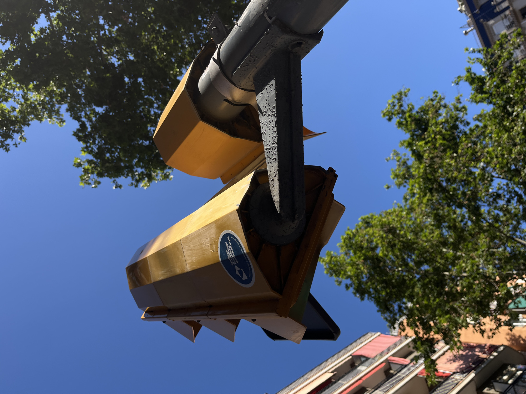 A low-angle shot focuses on an overhead pedestrian traffic signal in a city setting, likely during daylight hours. The signal, a light yellow with a black mount, is attached to a gray, cylindrical pole. The signal's lower portion includes downward-pointing triangular protrusions. A sticker is visible on the signal's side, bearing text in a language other than English. Clear blue sky fills the background.
Green-leafed trees partially obscure the view, with treetops appearing on the left and right sides. Portions of a multistory building, with light-colored walls and alternating red-and-white awnings, appear on the lower right. The location is Unknown, Unknown.