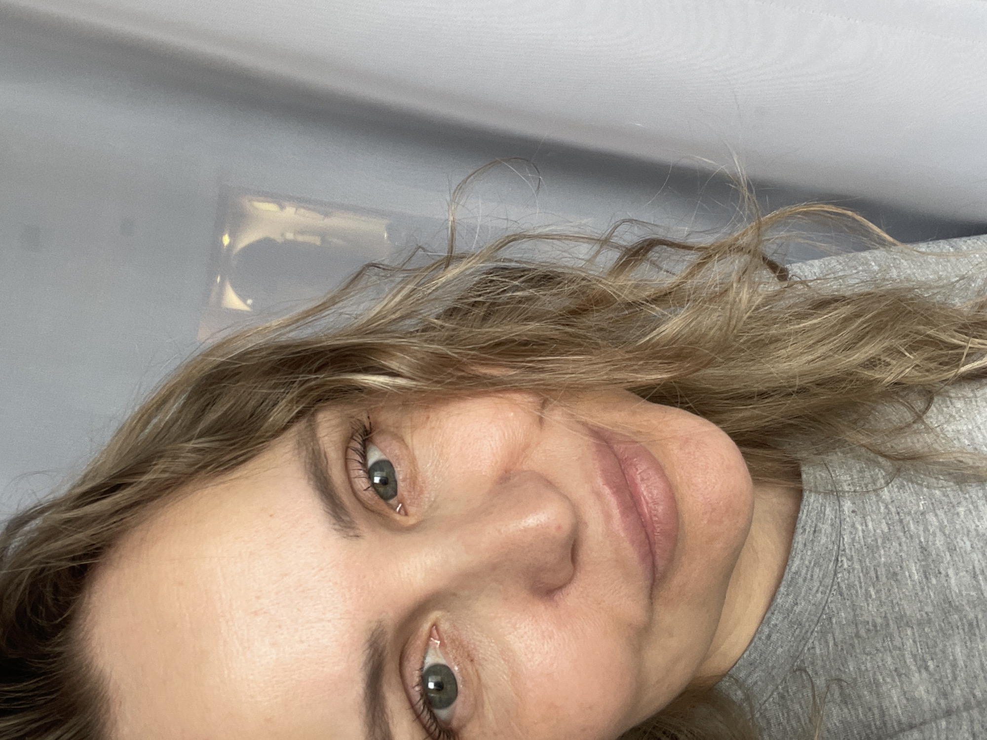 This media file presents an upright close-up portrait of a woman, rotated 90 degrees clockwise. She has long, wavy hair that is a mix of blonde and light brown tones. Her eyes are a bluish-gray color, and she is looking directly at the camera with a soft, slight smile, conveying a calm and friendly emotion. Visible eyelashes suggest the use of mascara. She is wearing a plain gray t-shirt, the textured fabric of which is visible on her shoulder and chest. The background is a simple, light gray surface, likely an indoor wall or ceiling, with a subtle, blurry reflection or light source appearing in the upper right portion of the image. There are no specific location cues or visible text within the frame, indicating a generic indoor setting. The lighting is even, illuminating her face clearly.