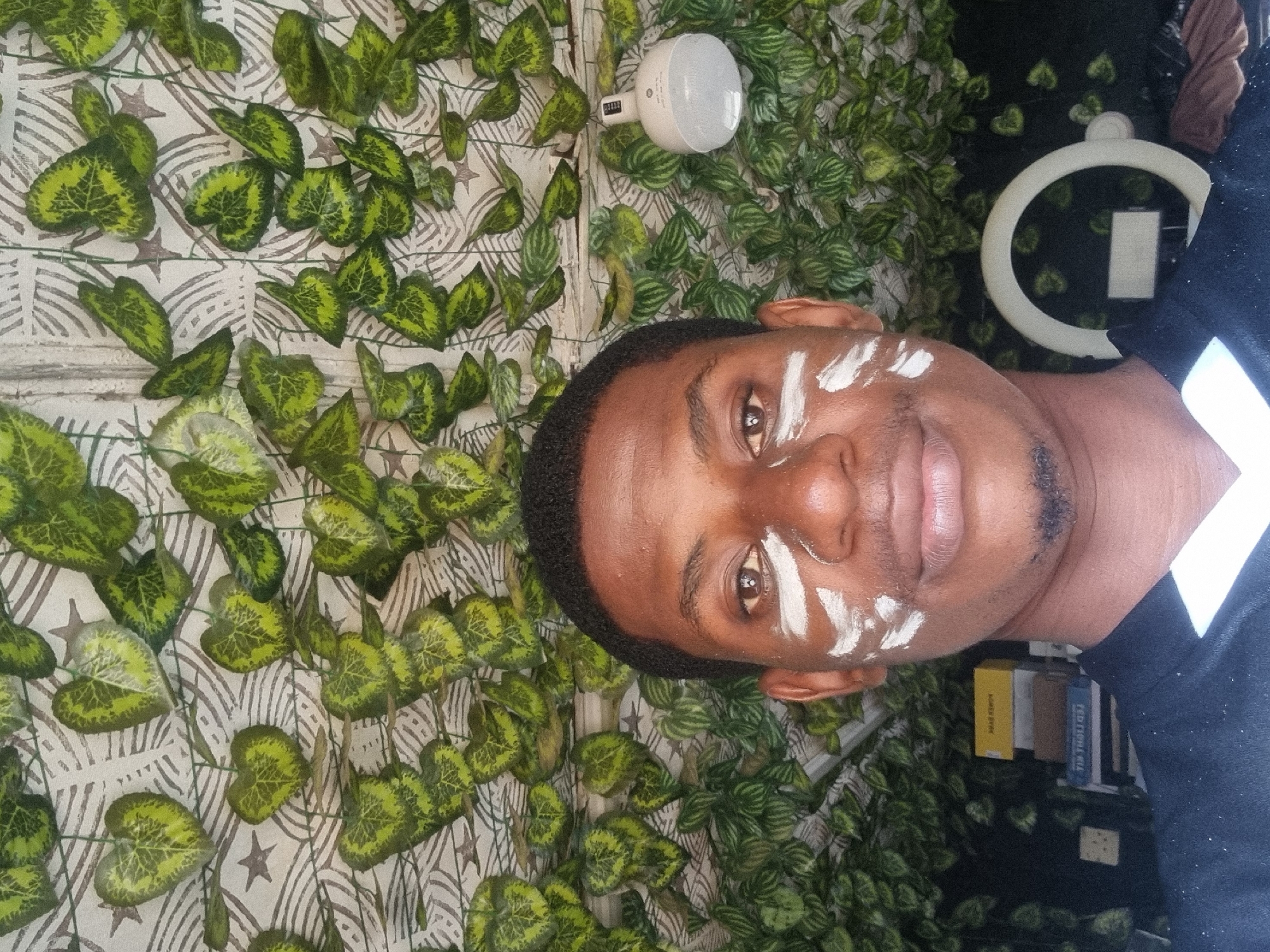 A close-up, slightly tilted portrait photograph shows a man with dark skin and short, dark hair. White markings are visible on his forehead and cheeks. He is wearing a dark-colored shirt with a white collar. Behind him is a backdrop of artificial greenery, resembling ivy, intertwined with a patterned wallpaper featuring stars and abstract designs. A white circular ring light is visible to the right of his head, and a white, cylindrical object is attached to the wall above his head. The background includes glimpses of books and a dark, possibly cluttered space. The location is plausibly Onitsha, Nigeria, but there are no definitive landmarks or cultural indicators to confirm this.