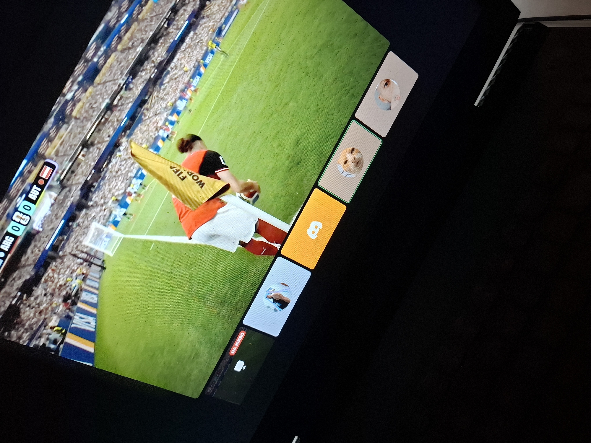An angled close-up shot captures a television screen displaying a soccer match. The score is visible at the top left: "ARG 0 - 0 AUT", indicating a game between Argentina and Austria. A player wearing an orange and white jersey is bending over on the green field, holding a yellow object with "FIFA WOR" printed on it. Below the main screen, a strip of smaller windows shows various content. From left to right, these include a live stream indicator with text in Cyrillic, a profile picture of a person, a Discord logo, a profile picture of a cat, and another profile picture of a person. The scene is dark outside the television screen, suggesting it's nighttime or an indoor setting. No specific location in Burgas, Bulgaria is discernible from the image.