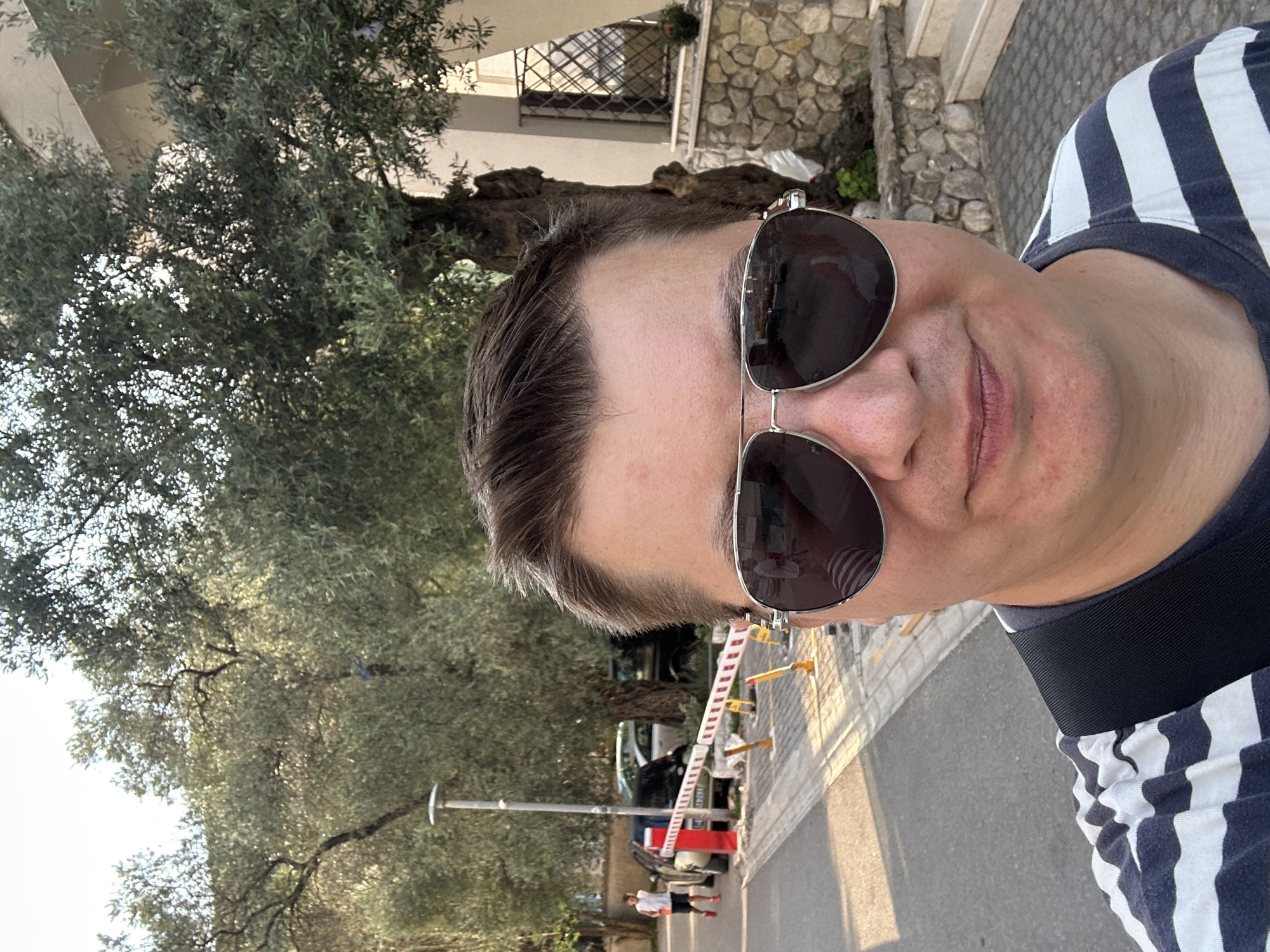 This is a selfie taken in Budva, Montenegro. The image is vertically oriented. The subject is a person wearing sunglasses and a navy and white striped t-shirt. They are looking at the camera, and their mouth is slightly open. The background shows an olive tree with dense foliage, a stone wall, and a building with a barred window. In the distance, there is a street with a barrier gate, a vehicle, and two people walking. The lighting suggests it is either early morning or late afternoon, with warm sunlight casting shadows. The overall mood appears casual and relaxed. There is no visible text in the image.