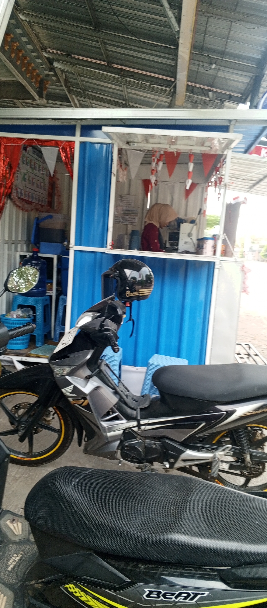 The image shows a scene at a roadside stall in Pingkuk, Indonesia. In the foreground, a black and silver motorcycle is parked. The motorcycle has a black seat and yellow accents on its wheels. A black helmet with a gold stripe is resting on the handlebars of the motorcycle. Behind the motorcycle, the stall structure is visible, with bright blue corrugated metal walls.
Inside the stall, a person wearing a light brown hijab and a maroon shirt is visible, appearing to be working at a counter with a laptop and a small appliance, possibly a cash register or a dispenser. The stall is decorated with red and white triangular pennant flags. To the left of the stall, blue plastic stools and water dispensers are stacked. The roof of the stall is made of corrugated metal, and the surrounding environment appears to be outdoors, with some foliage visible in the background. The ground appears to be paved or dirt.