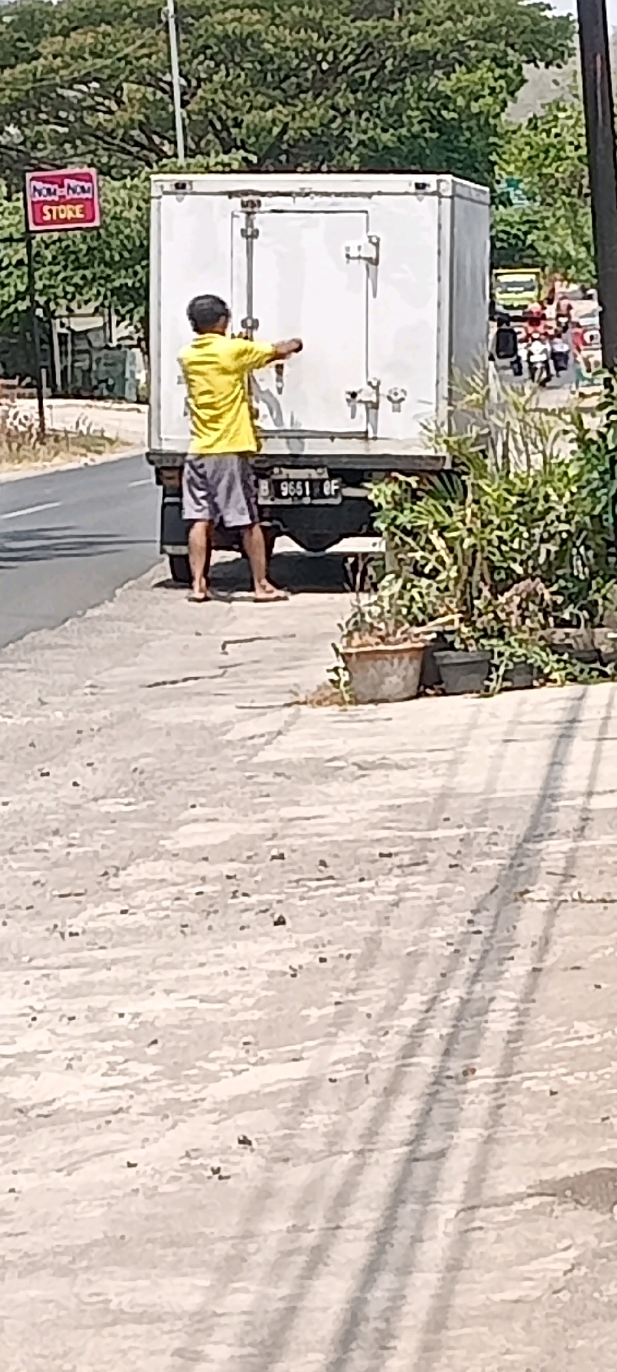 A man in a yellow shirt and gray shorts stands in front of a white truck with its rear doors open. He appears to be reaching into the truck. The truck has a license plate that reads "B 9651 OF". To the left of the truck is a sign that says "NOM-NOM STORE". In the background, there are trees and buildings. The scene is outdoors on a paved surface, possibly a street or parking lot. The ground is light-colored with some scattered debris. The image appears to have been taken during daylight. The location is identified as the city of Bulu, Indonesia.