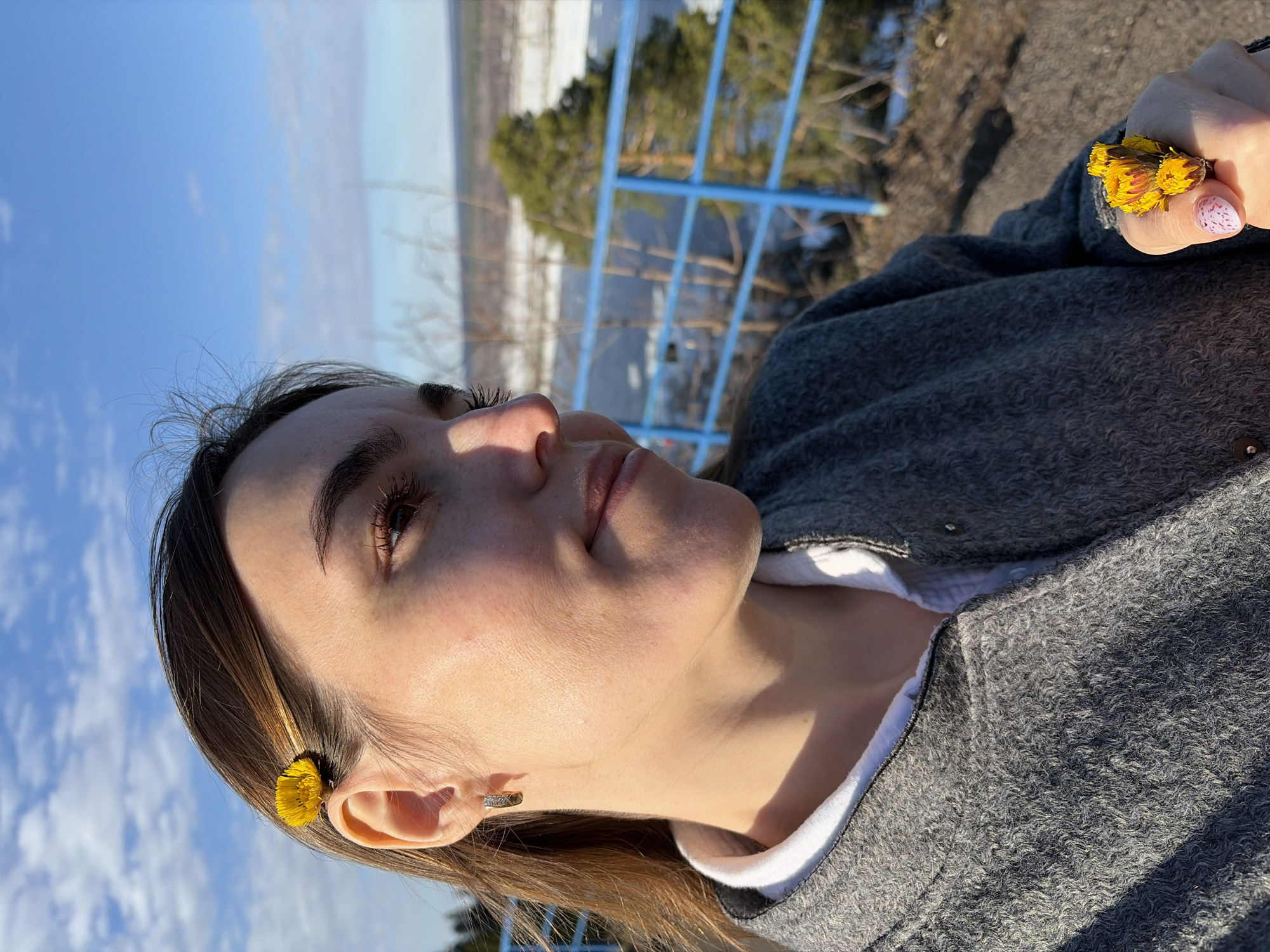 The image shows a young woman outdoors, likely in a natural area within the city of Sarapul, Russia. She is wearing a gray jacket over a white top. She has a yellow flower in her hair near her ear, and is holding a cluster of yellow flowers in her hand. Her fingernails are painted with a patterned design. In the background, a blue railing can be seen, with trees and a landscape visible beyond it. The sky is blue with some clouds.