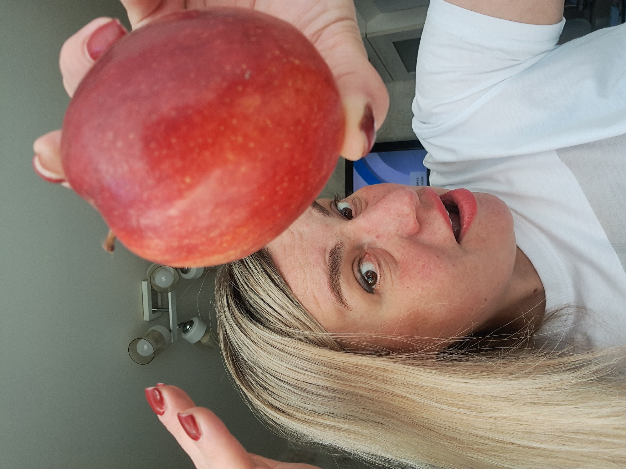 A woman with blond hair holds a red apple up to the camera. She is wearing a white t-shirt and has red nail polish. The background is out of focus, but shows a wall and a light fixture.
Мининская Поляна Кп, Russia is not discernible.