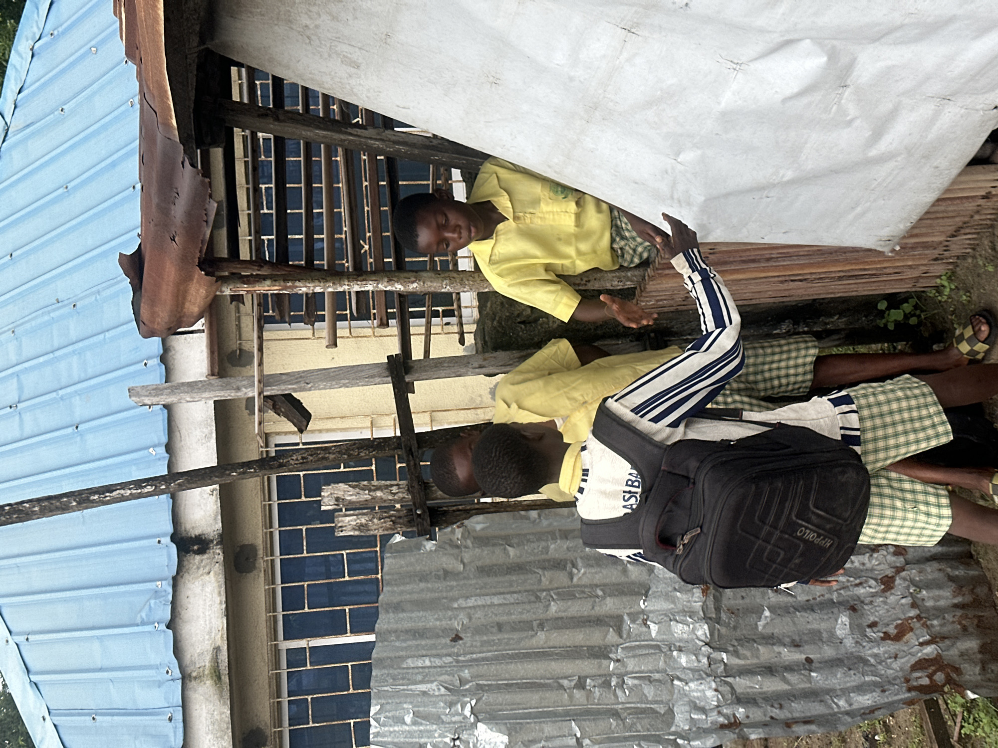 The image depicts three young boys dressed in school uniforms, gathered in an outdoor setting that appears to be the city of Badagry, Nigeria. Two of the boys, wearing yellow shirts and checkered shorts, are interacting within a makeshift structure constructed from wood and corrugated metal. One of these boys is gesturing with his hands as if speaking. The third boy, in the foreground, has a black backpack on and is facing away from the camera, his striped shirt and checkered shorts also indicating a school uniform. The environment shows signs of disrepair, with a corrugated metal wall and a rusted tin roof over the structure. A building with blue-paned windows and a light-colored facade is visible in the background, beneath a blue metal roof. The ground is uneven and appears to be dirt with some sparse vegetation.