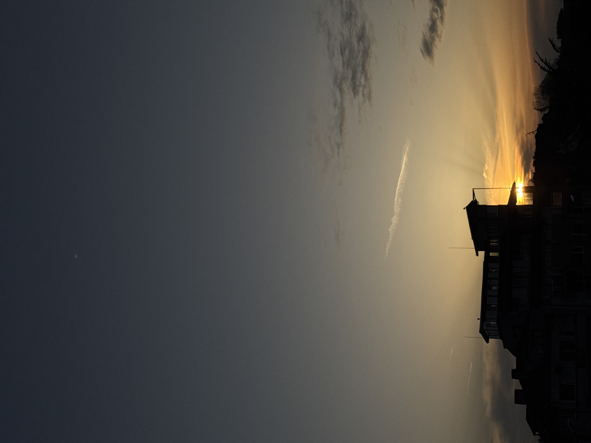 The image shows a building silhouetted against a sunset. The sky is dusky with rays of light extending out of the setting sun. There are clouds present, some darker than others, and there are also contrails present. The location in Tsarevo, Bulgaria, is not determinable from the image.