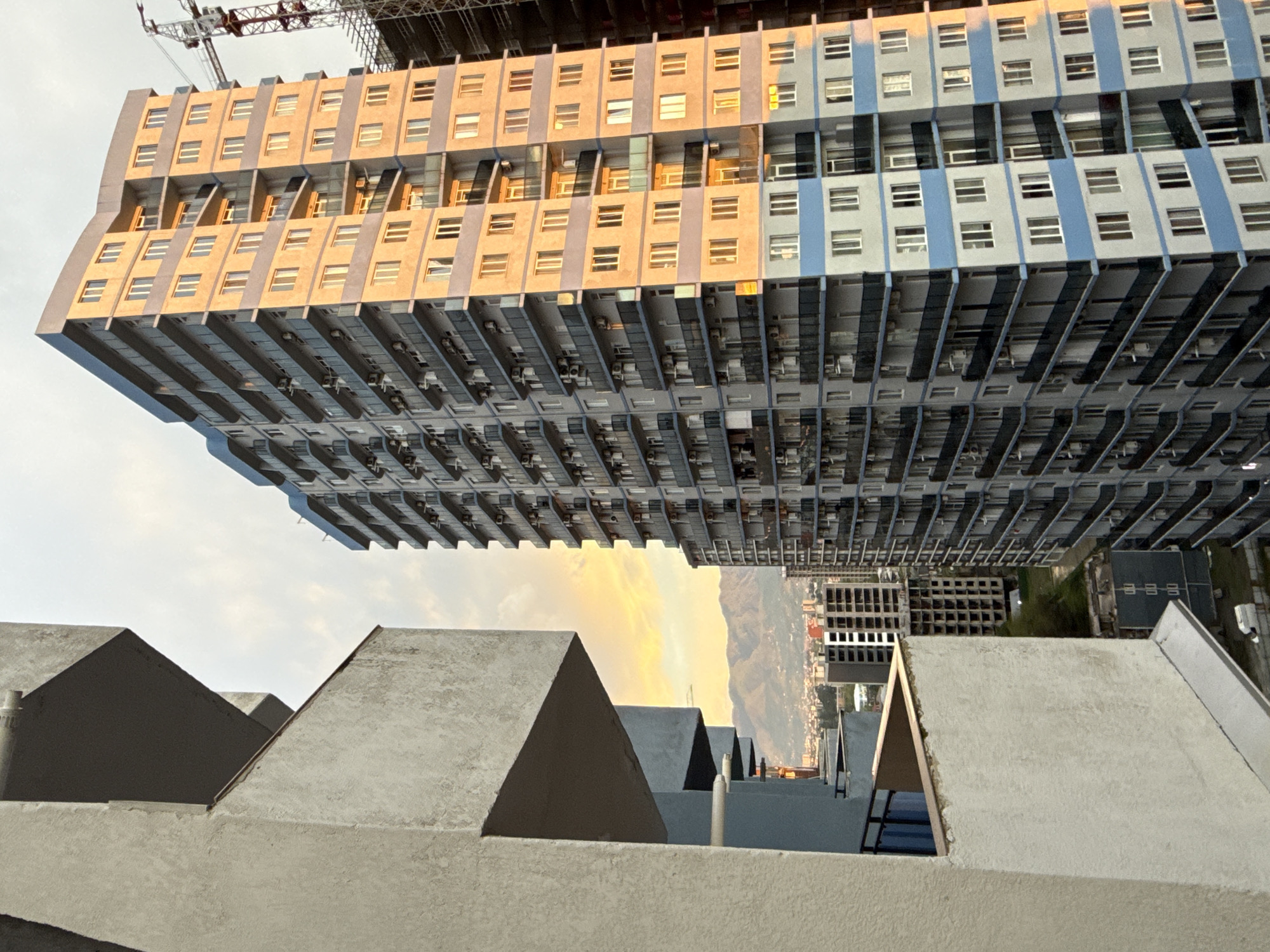 The image shows an upward perspective of a large, multi-story building under construction in Batumi, Georgia. The building's facade is comprised of horizontally stacked floors with rows of windows and air conditioning units. The color scheme alternates between yellow and blue hues. A construction crane is visible atop the building. In the foreground are several gray, triangular-shaped structures, possibly rooftops. Between these structures, a distant cityscape is partially visible with mountains and a glimpse of a sunset.