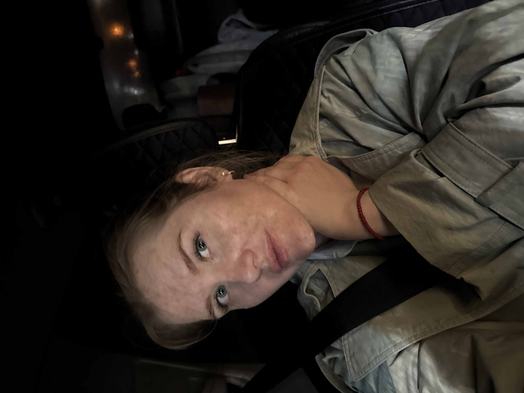 A young woman is lying down, presumably in a car, wearing a light-colored jacket and a red bracelet on her wrist. A black seatbelt strap is visible across her body. The lighting is dim. The location is Горный, Russia.