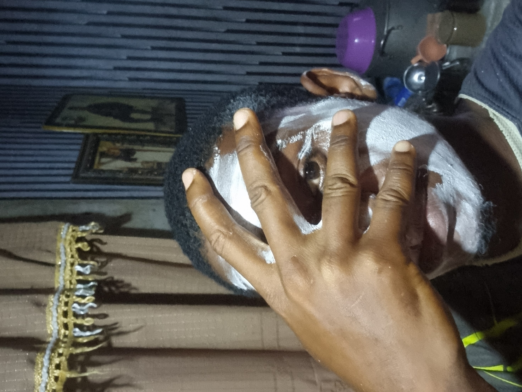The image presents a close-up, angled view of a person with dark skin in an indoor setting, likely in Onitsha, Nigeria. The central subject is the person's head and right hand. A thick layer of white substance, possibly chalk or paste, is applied to significant portions of their face. One eye is distinctly visible, featuring a dark iris and clear white sclera, looking directly towards the camera. The person's right hand, also with dark skin, is placed across their face, fingers spread, appearing to be part of the application process or covering part of the face. The person has short, dark, curly hair, and a dark garment with yellow/lime green accents is visible on their upper body.
The background suggests a domestic environment. On the left, two framed pictures are stacked on a surface, though their images are indistinct. In the upper right, various household items are visible, including a purple bowl, a silver pot or cup with a 'G' symbol, and other containers, indicating a possible kitchen or utility space. A decorative item with intricate gold and silver patterns is seen on a horizontally striped surface in the lower left. The wall behind the subject features a dark, striped texture. The lighting is artificial and somewhat dim, highlighting the direct gaze of the visible eye and the facial application.