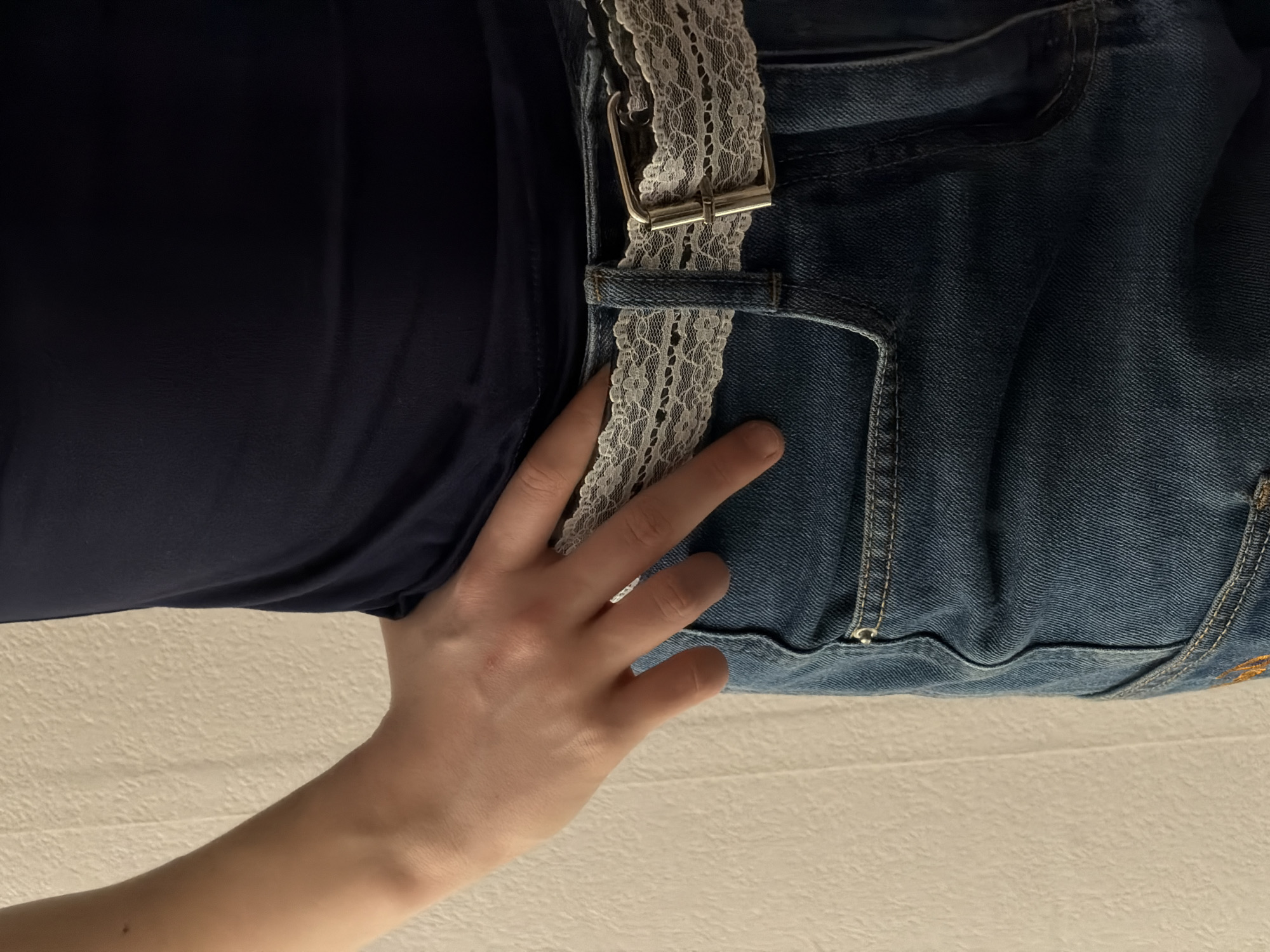 The image shows a person wearing blue jeans with a lace belt and a dark blue top. A hand with a ring on the finger is resting on the jeans near the belt. The background is a plain, light-colored surface, possibly a bed or table. There is no visible evidence indicating the location of Perm, Russia.