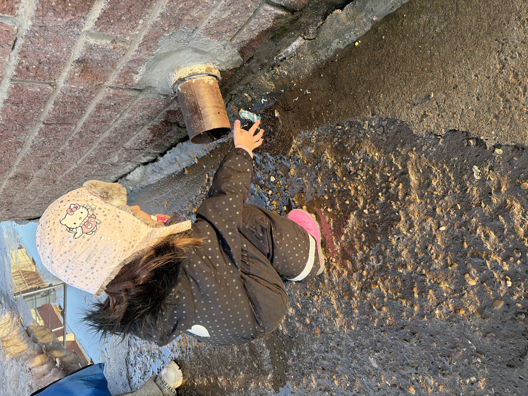 A young child is observed squatting on damp, uneven concrete ground adjacent to a reddish-brown brick wall in Chaikovsky, Russia. The child, viewed from the rear-right, wears a light pink winter hat adorned with silver stars and a "Hello Kitty" emblem, a dark-colored winter jacket with light polka dots and a white heart detail, dark pants, and a bright pink boot on the right foot. Their right hand is extended into a shallow puddle containing a small, crumpled blue and white object, possibly a plastic wrapper, and other debris. A rusted metal pipe protrudes horizontally from the lower section of the brick wall, secured by grey cement, with water appearing to emanate from the area, contributing to the wet ground and a narrow stream flowing along the wall's base. Sunlight illuminates parts of the wet ground, creating reflections. To the far left, a portion of another individual's leg and a light-colored shoe are visible near a larger puddle, which reflects an indistinct architectural structure.