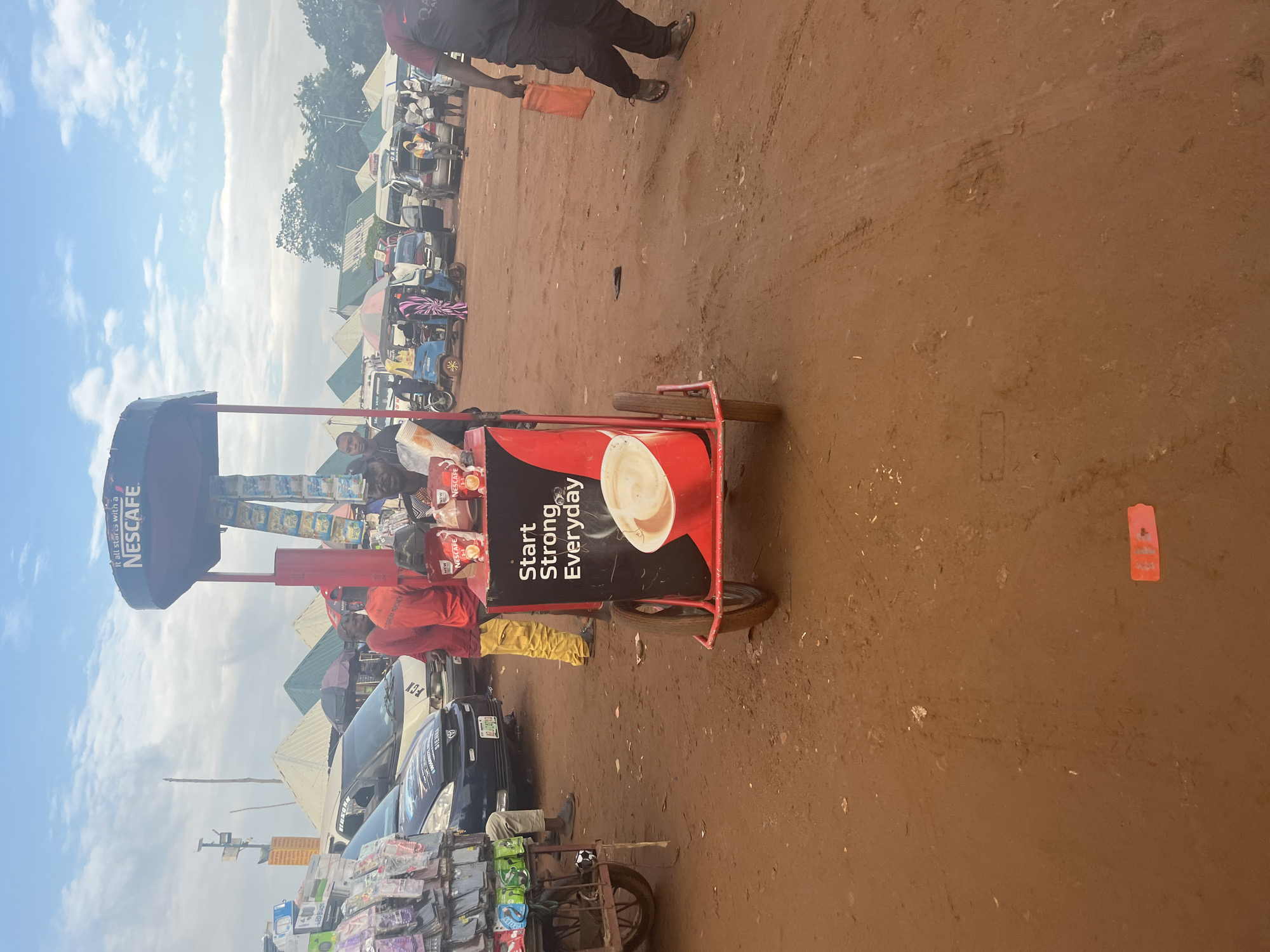 This is a medium shot of a street market scene in Nyanya, Nigeria, under a partly cloudy sky. In the foreground, a red and black mobile NESCAFÉ beverage cart is prominent, displaying the slogan "Start Strong Everyday" and images of coffee. Several NESCAFÉ packets are stacked on top of the cart. To the left of the cart, a stall displays various small electronic accessories on racks.
Behind the cart, several people are visible. Two men in red shirts and yellow trousers stand near the cart. Further back, various vehicles, including a white van and a dark sedan, are parked. Tents are set up in the background, suggesting a marketplace. A man in a dark t-shirt and black trousers is walking away from the cart towards the right side of the frame, carrying an orange bag. The ground is composed of reddish-brown dirt, with tire tracks and scattered debris.