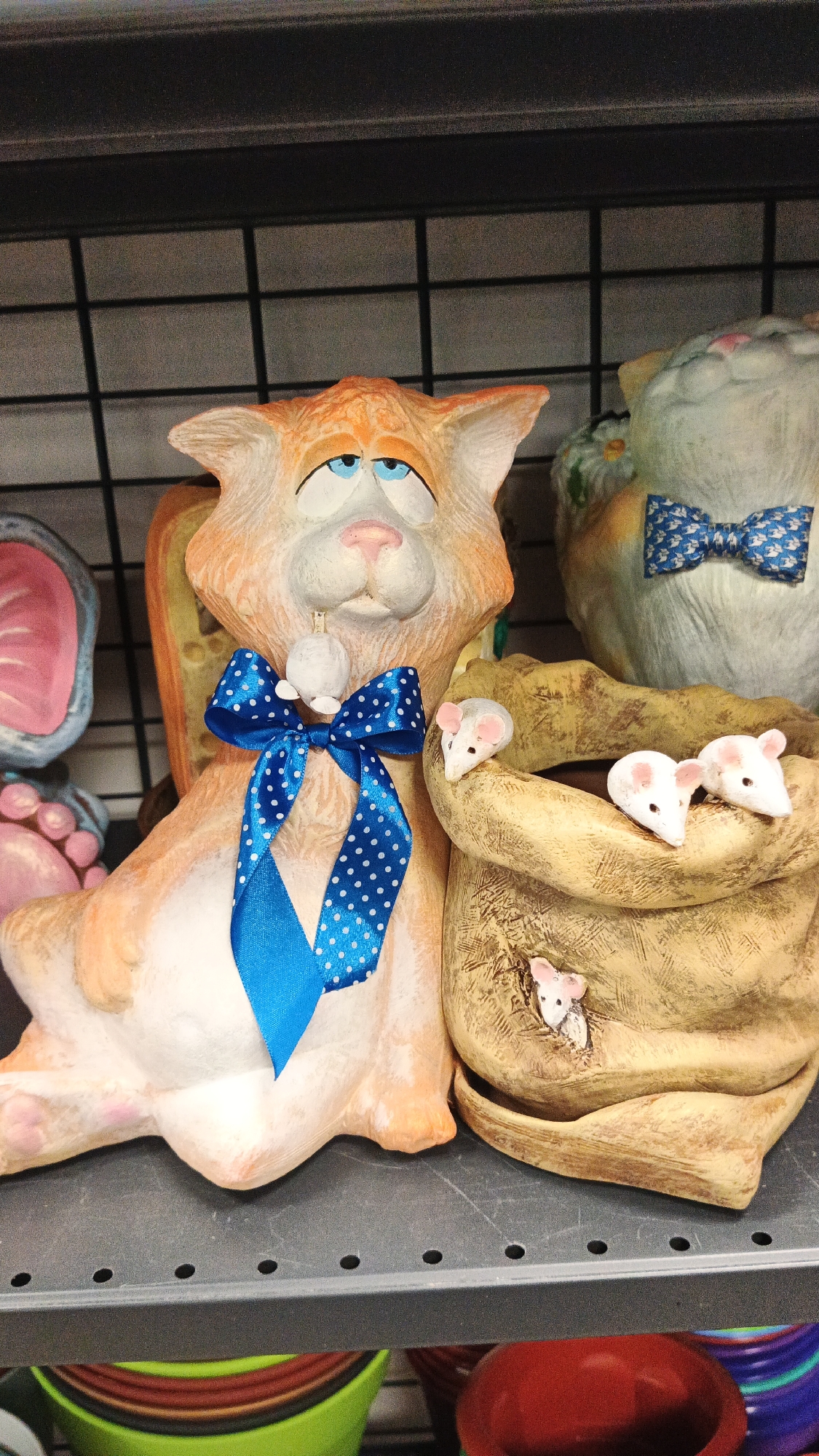 The image depicts a shelf in a store, likely in Kryvyi Rih, Ukraine, displaying various ceramic decorative items. The primary focus is on two ceramic cat figures. The left figure is a large, orange tabby cat with blue eyes, a pink nose, and a sad expression. It is adorned with a blue satin ribbon with white polka dots tied around its neck, and a small white mouse figurine is attached to the ribbon. Behind the orange cat is a slice of cheese-shaped ceramic. To the right of the orange cat is a tan, sack-shaped ceramic pot. Several small white ceramic mice with pink ears are positioned on and around the rim of the sack. A second, larger ceramic cat figure is visible behind the sack, partially obscured, with a blue and white patterned bow tie. The background shows a black wire shelf and other indistinct items on shelves below. The overall scene is static and appears to be a retail display.